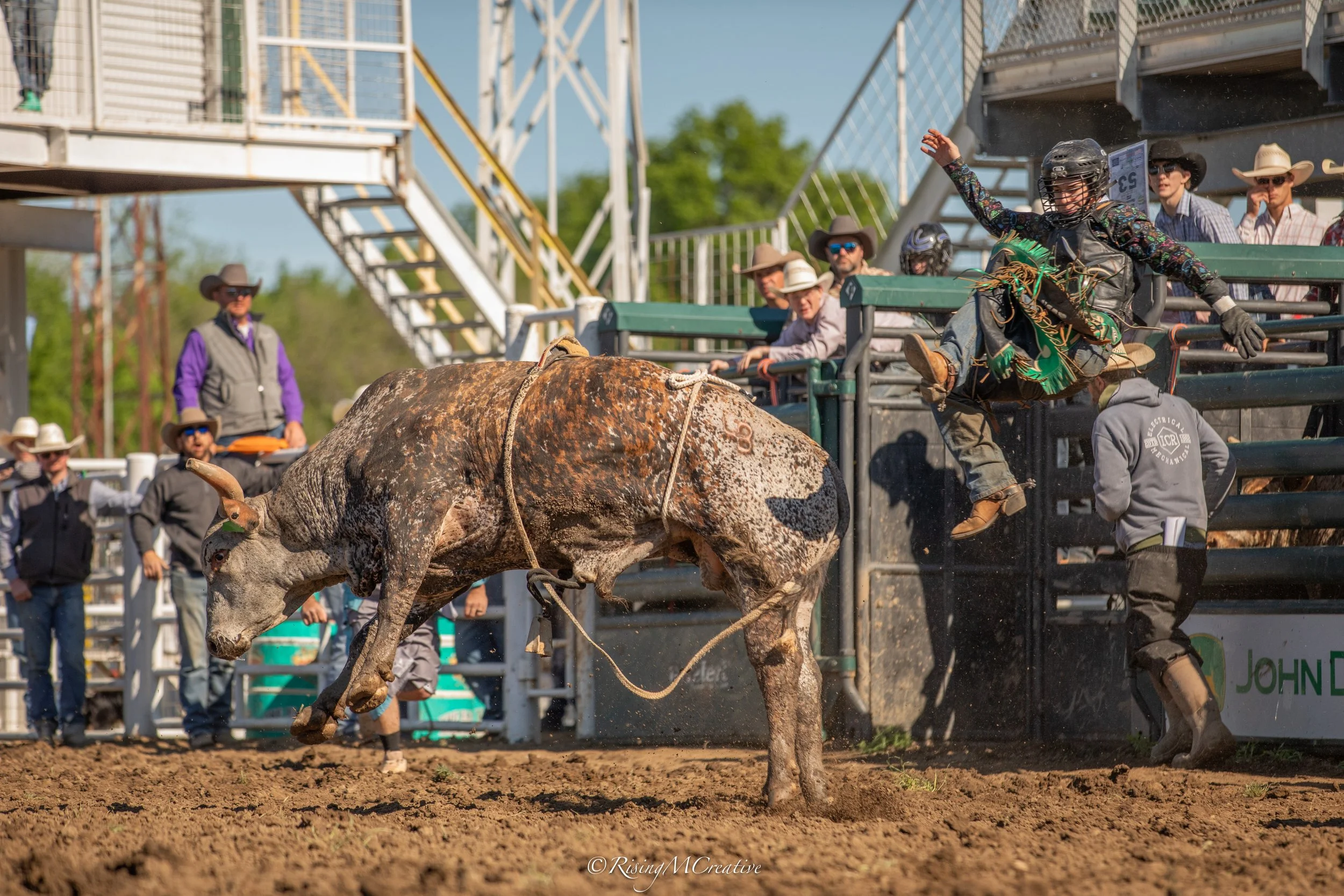 A rodeo scene with a bull rider in a black helmet and vibrant shirt landing on the ground after attempting to stay on a bucking bull, with spectators and rodeo officials watching in the background under a clear blue sky.