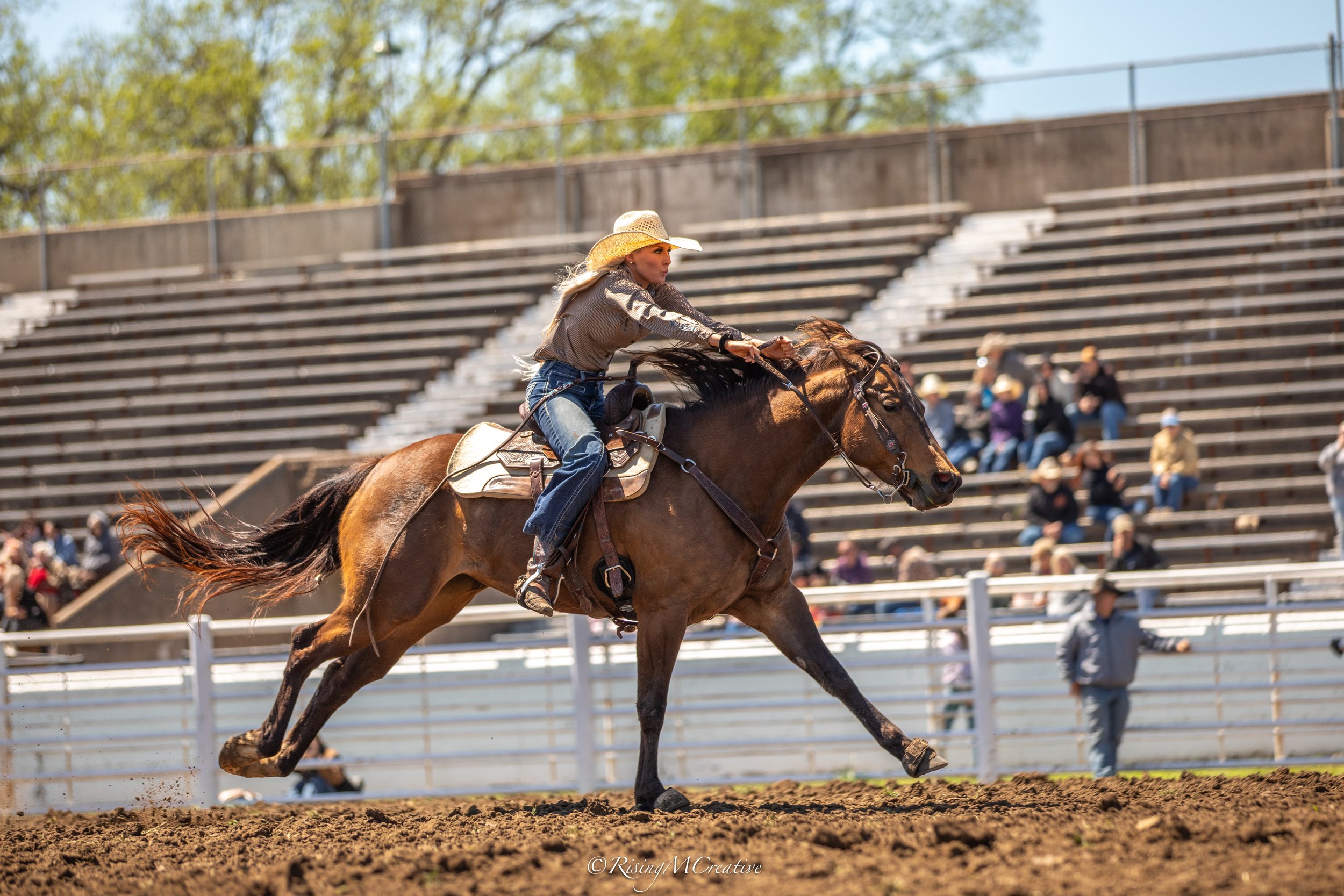 A woman riding a horse in a rodeo or equestrian event, wearing a cowboy hat and western attire, with spectators watching from the bleachers in the background.
