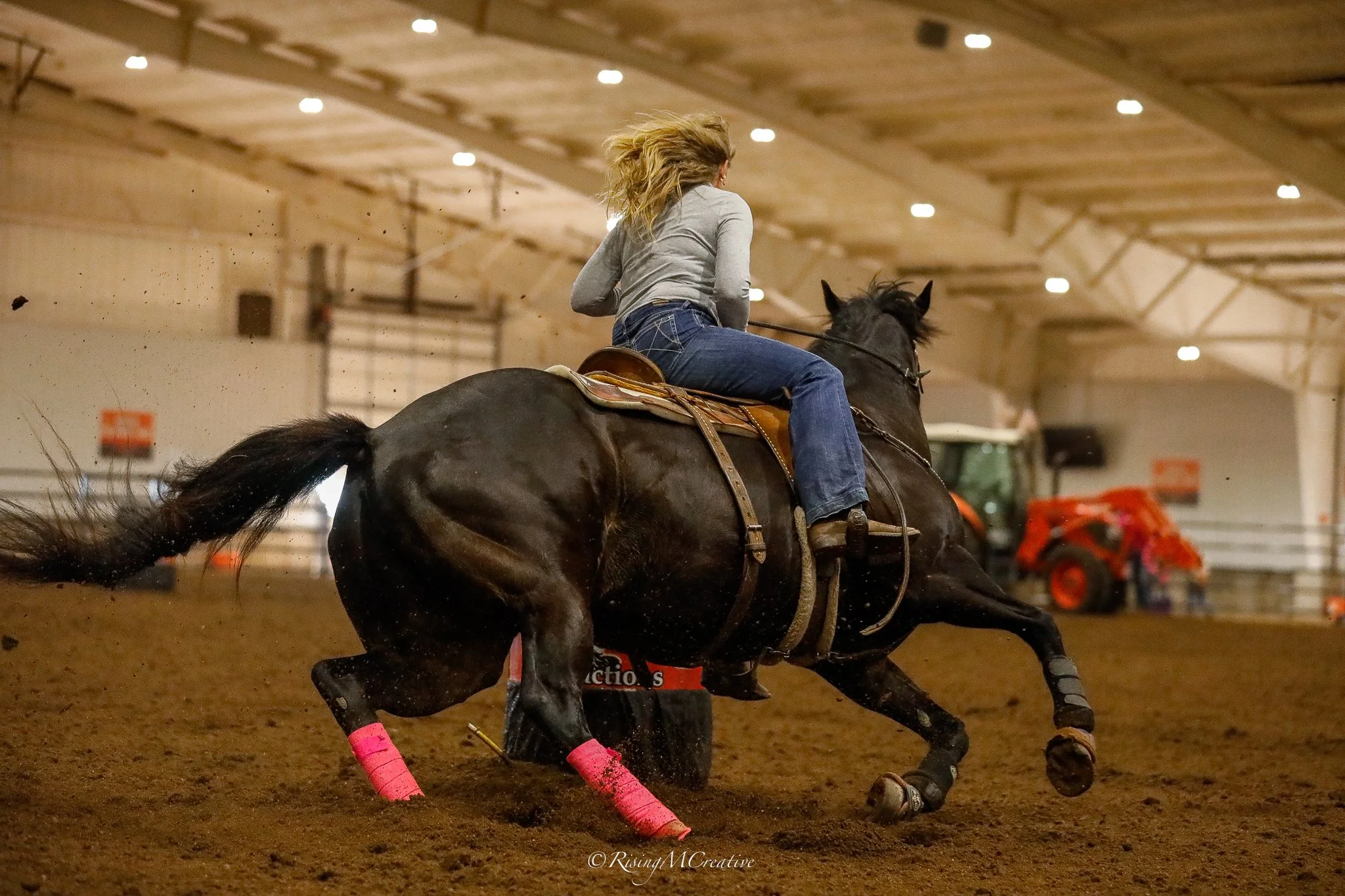 A person riding a horse and racing around an indoor arena, wearing gray and jeans, with pink leg wraps on the horse, kicking up dirt as they turn.