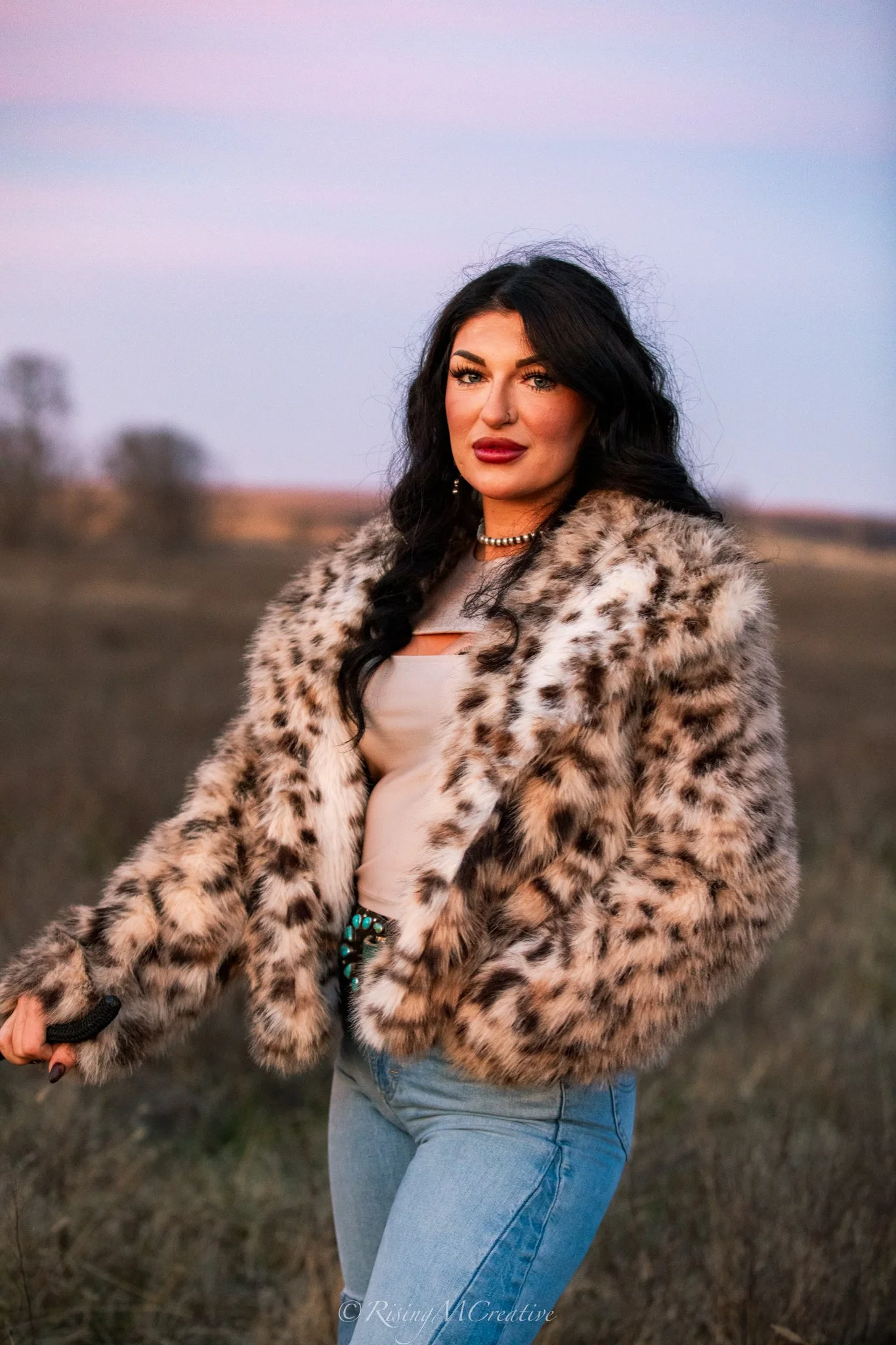 A woman with black hair and makeup wearing a leopard print fur coat, beige top, blue jeans, and jewelry standing outdoors at sunset or dusk on a field.