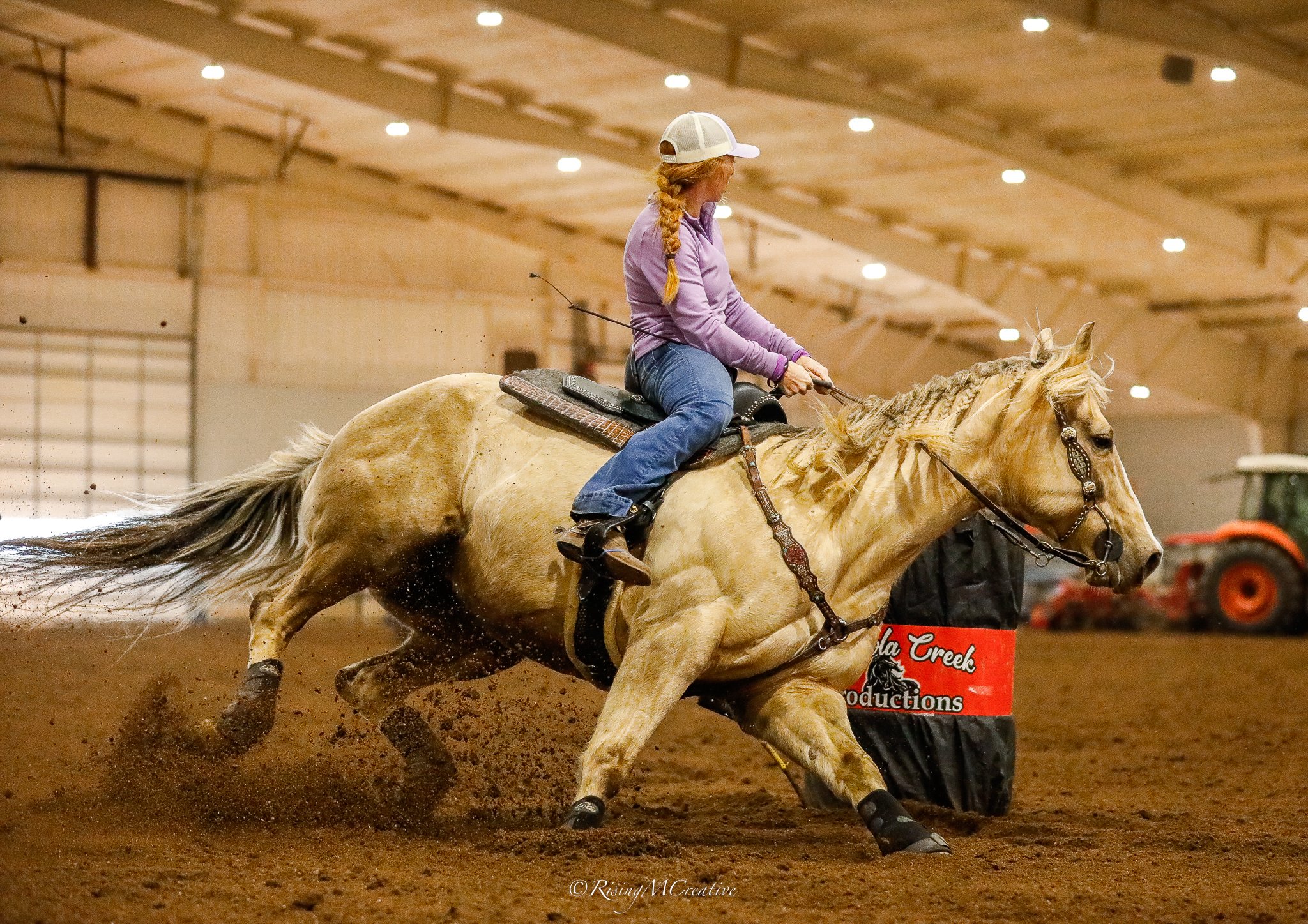 A woman riding a palomino horse during an indoor equestrian event, wearing a white cap and purple jacket, with a sign that reads 'Tala Creek Productions' in the background.