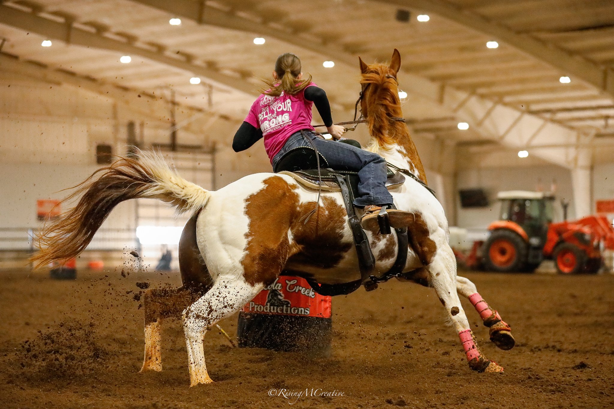 A person riding a pinto paint horse with a pink saddle pad inside an indoor arena, kicking up dust while performing a riding maneuver.