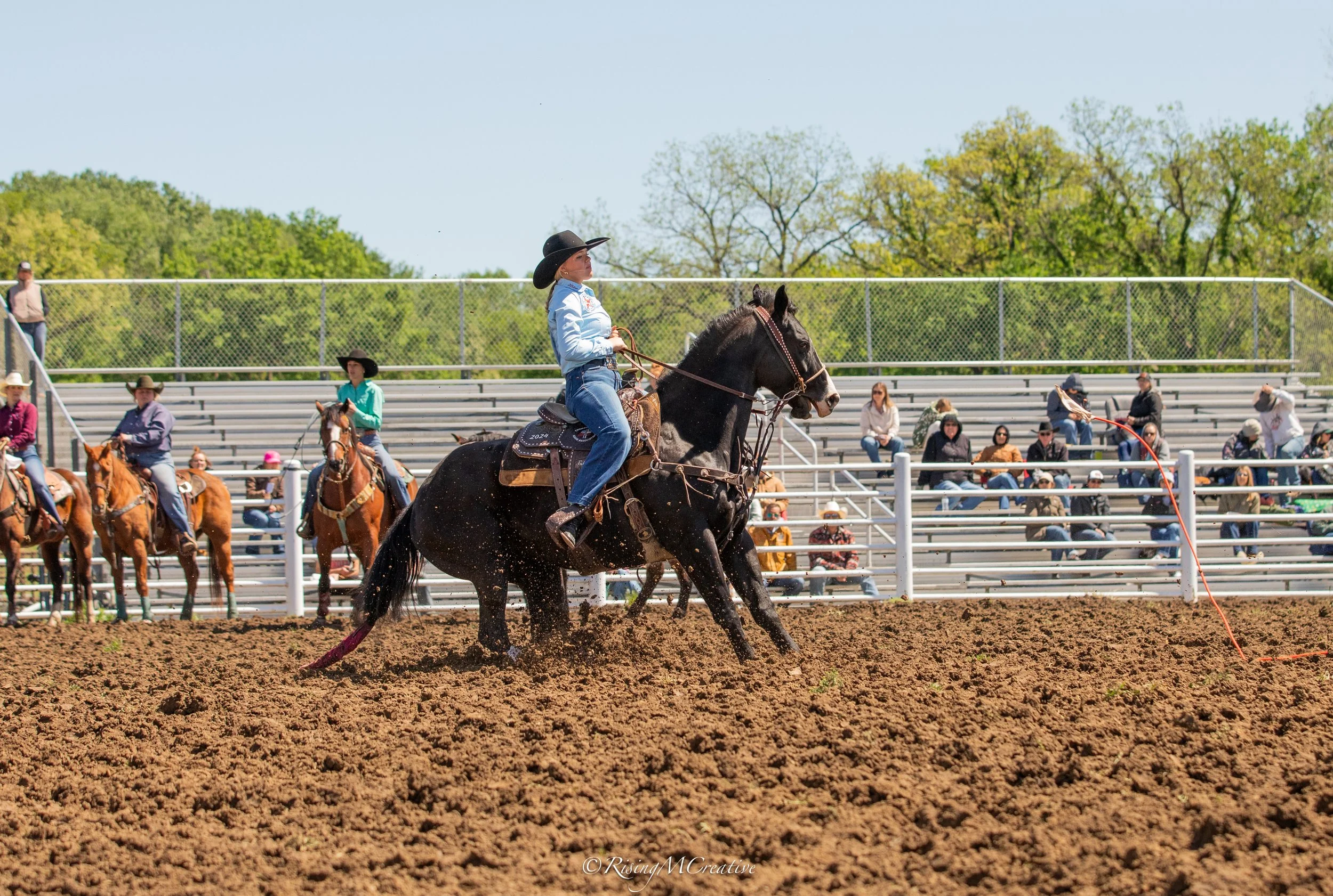 A woman wearing a cowboy hat rides a bucking horse during a rodeo event, with spectators seated on bleachers and trees in the background under a clear blue sky.
