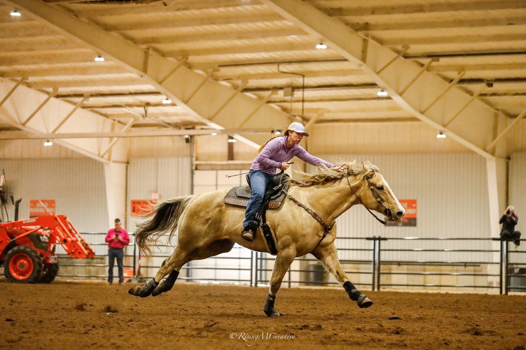 A woman in a purple jacket and white helmet riding a light-colored horse in an indoor arena. She is leaning forward holding the reins, with horses and people visible in the background.