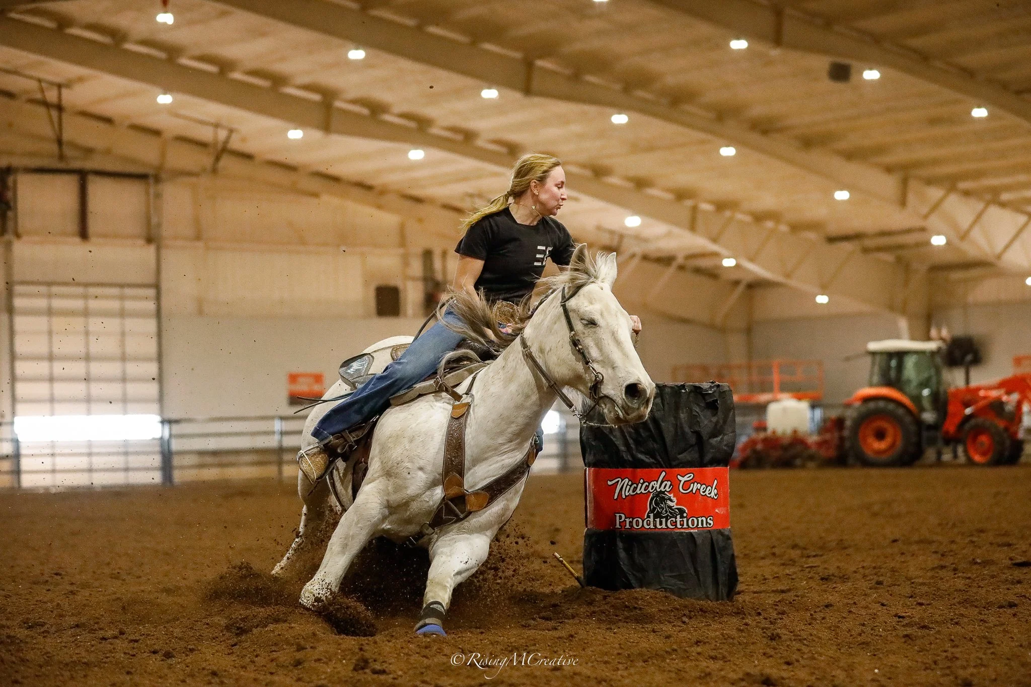 A woman riding a white horse in an indoor arena, participating in a barrel racing event. There are orange tractors and farming equipment in the background.