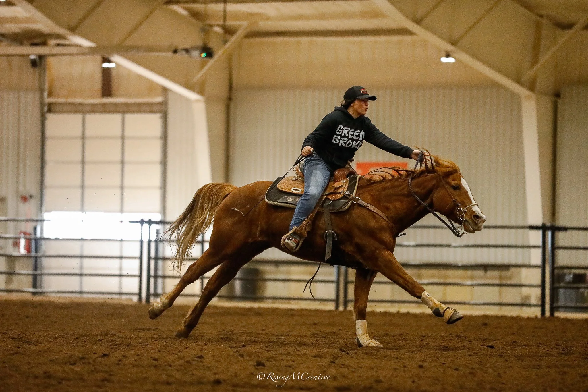 A person riding a galloping horse inside an indoor arena, wearing a black hoodie and cap.