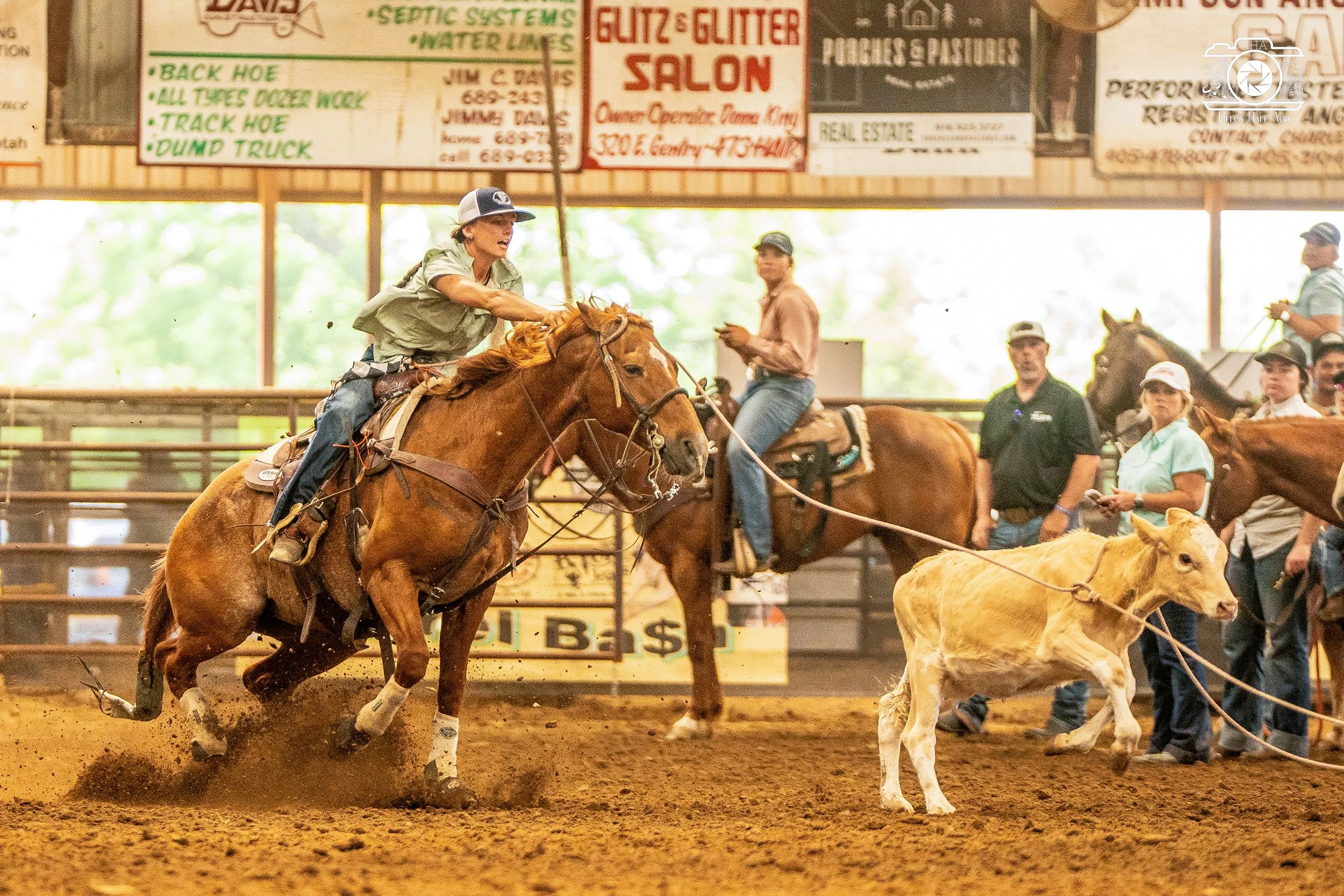Rodeo scene with a cowboy on a bucking horse trying to stay mounted as a calf is tied to the horse's reins; spectators and other riders are in the background inside an indoor arena with billboards overhead.