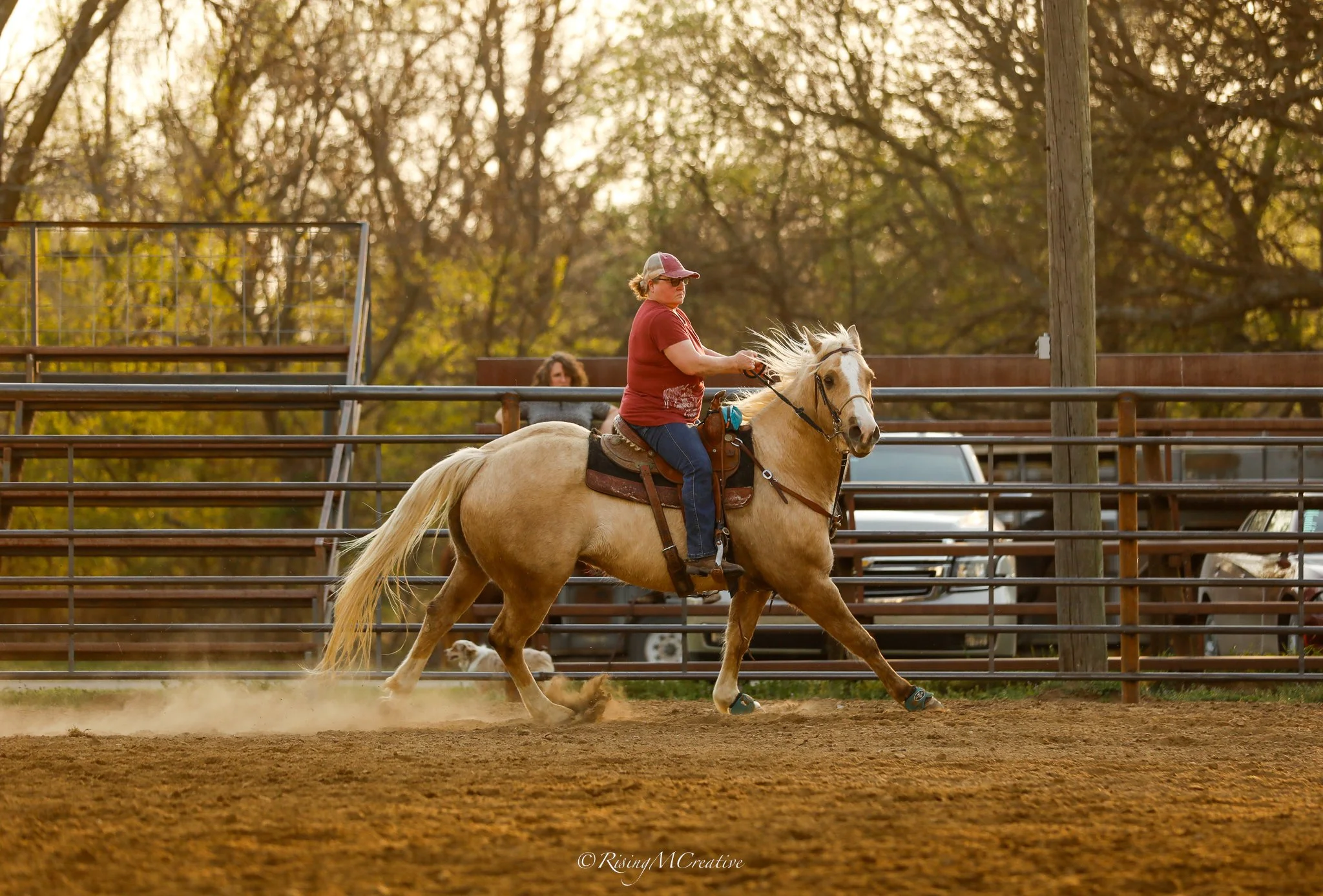 A woman wearing sunglasses and a baseball cap riding a galloping light-colored horse in an outdoor arena during sunset, with trees and a wooden fence in the background.
