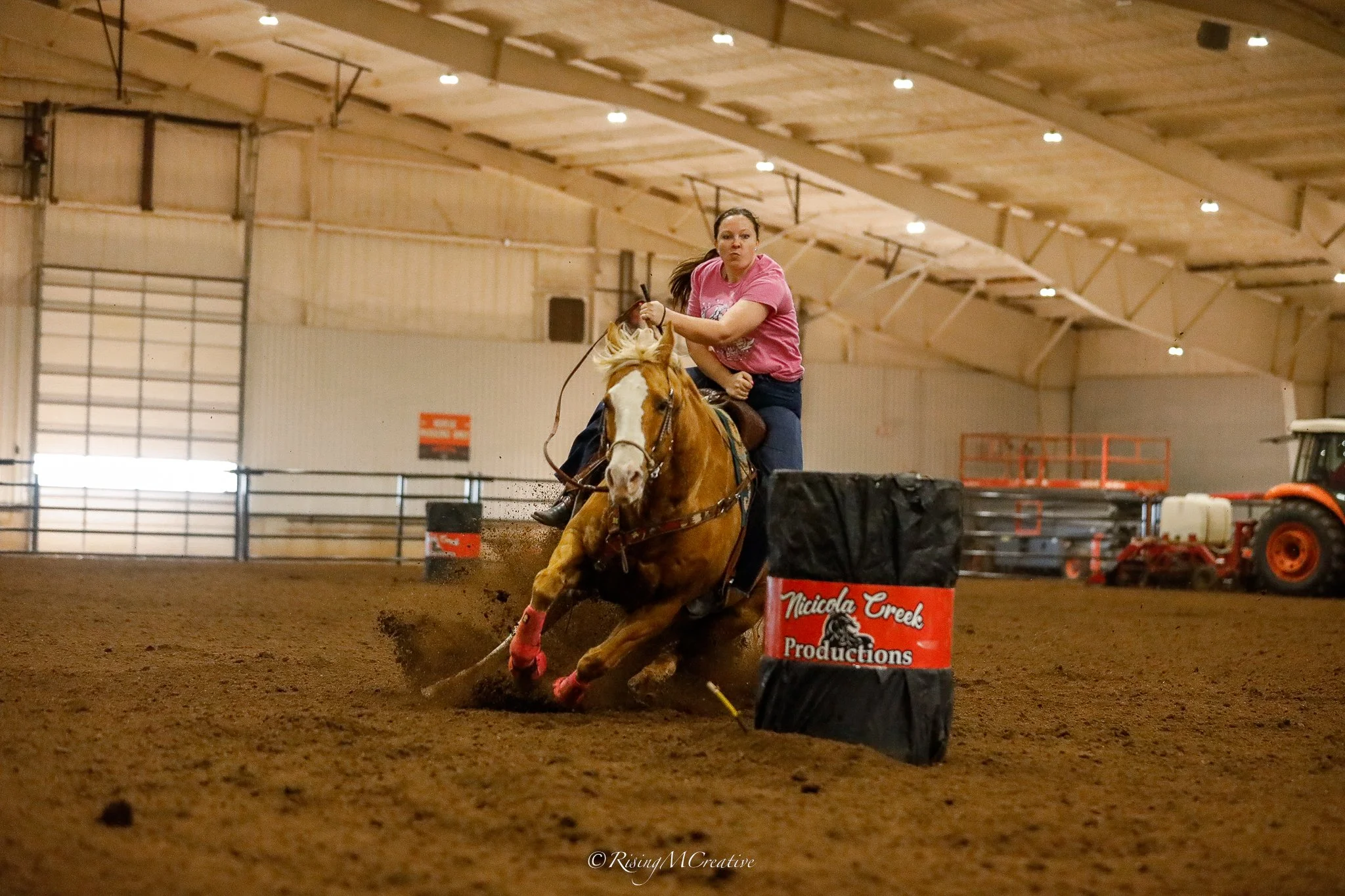 A woman riding a horse in an indoor arena during a barrel racing event. The horse is turning sharply around a barrel marked with 'Nicci Creek Productions.' The woman appears focused as she guides the horse through the course.