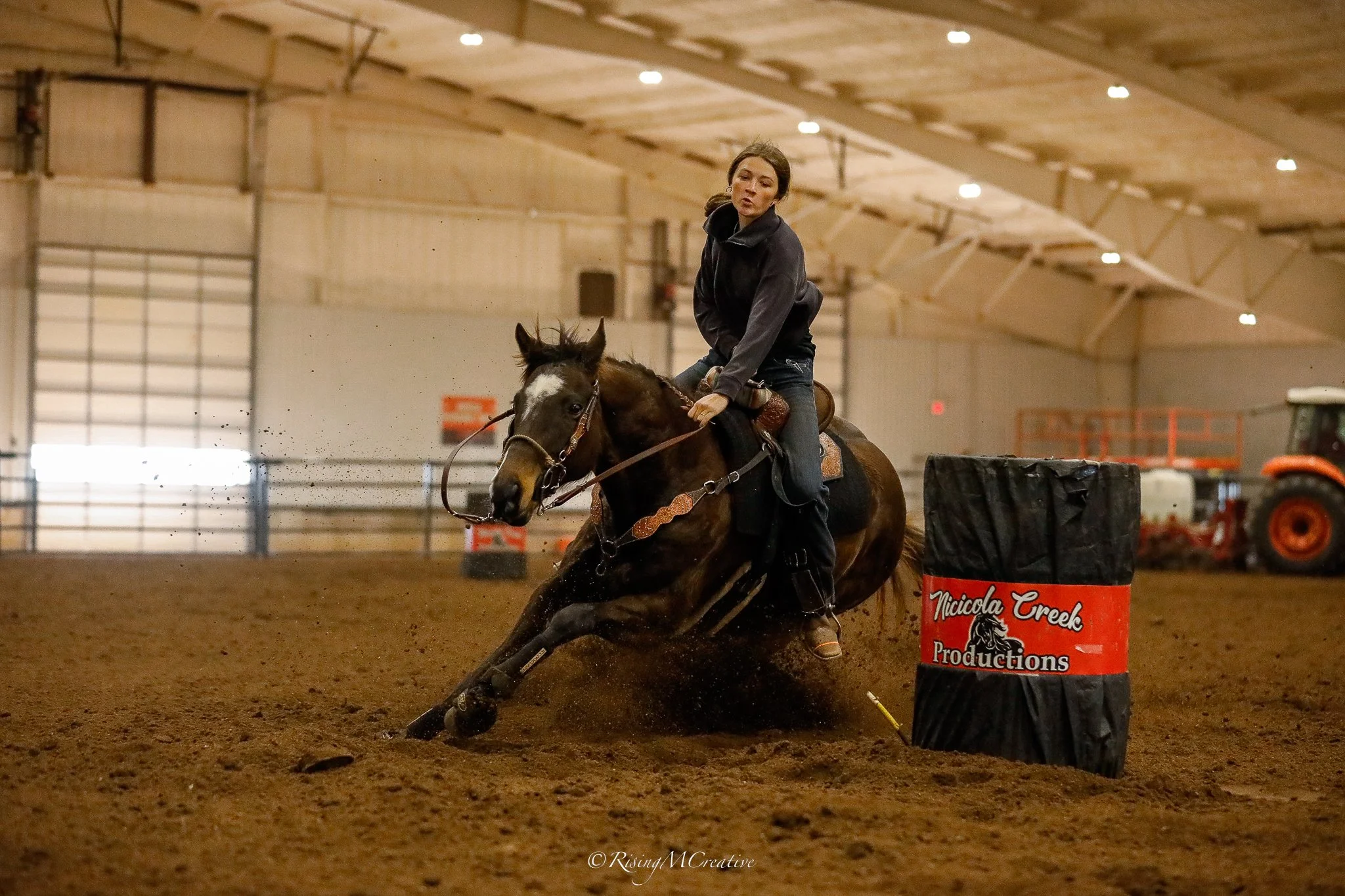A woman riding a horse during an indoor barrel racing event, navigating around a barrel marked 'Nicola Creek Productions'.