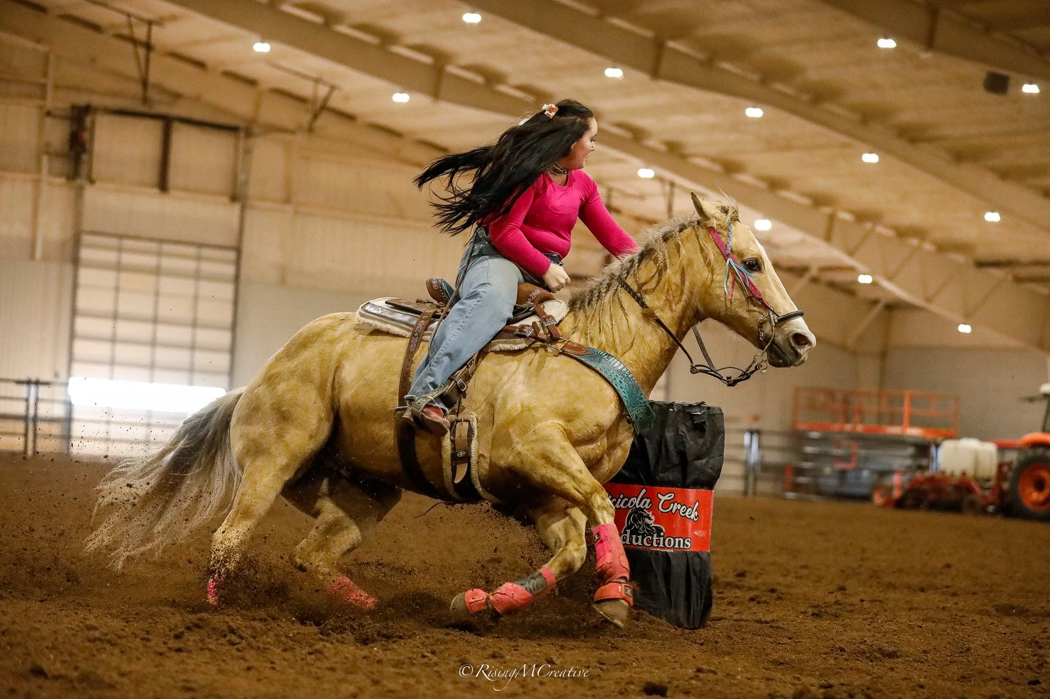A female rider with long dark hair, wearing a pink top and light pants, riding a palomino horse with a braided mane inside an indoor riding arena. The horse and rider are in motion, kicking up dirt as they navigate around a obstacle.