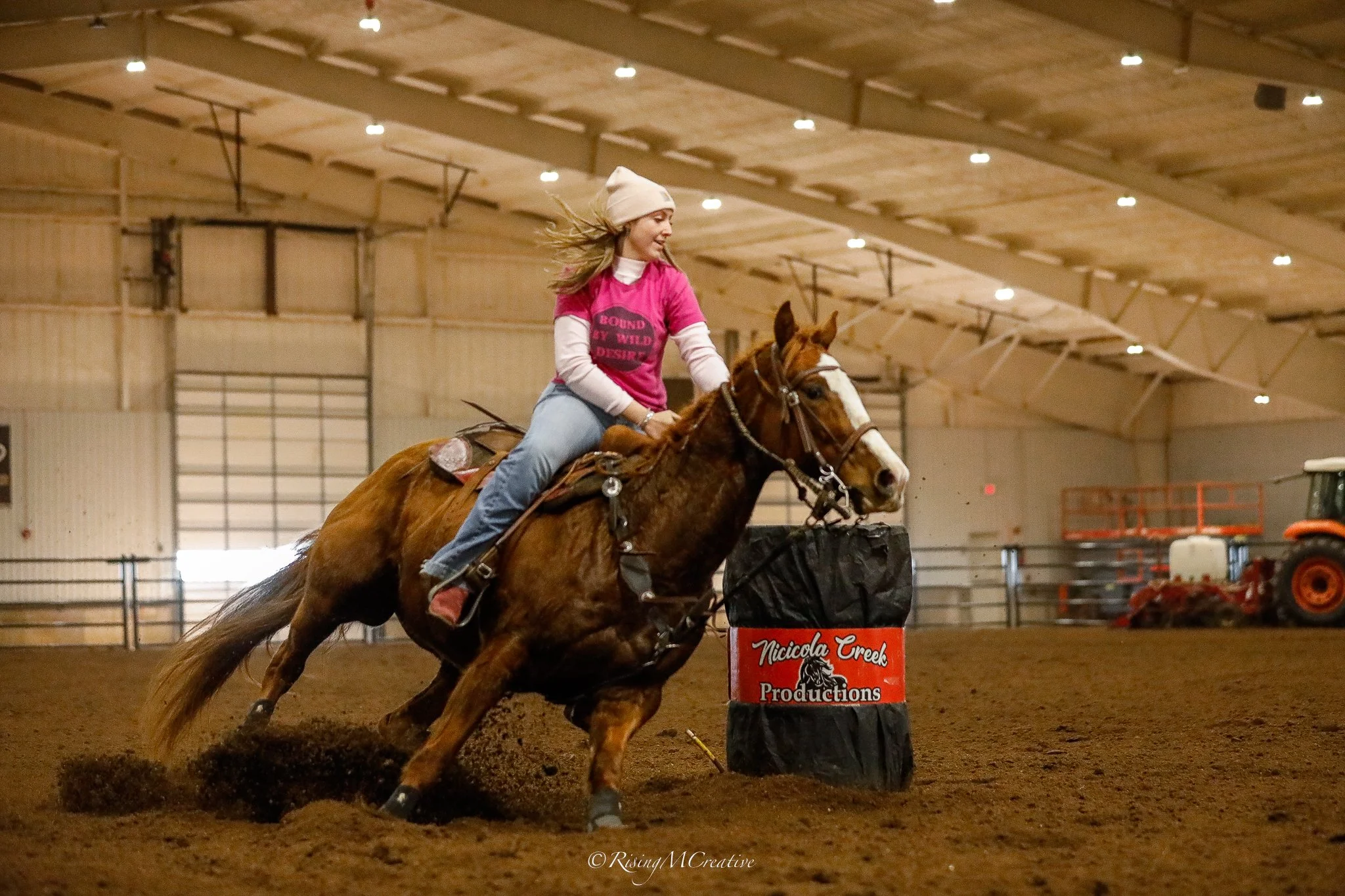 A young woman in a pink shirt and white hat rides a galloping horse inside an indoor arena, participating in a rodeo event.