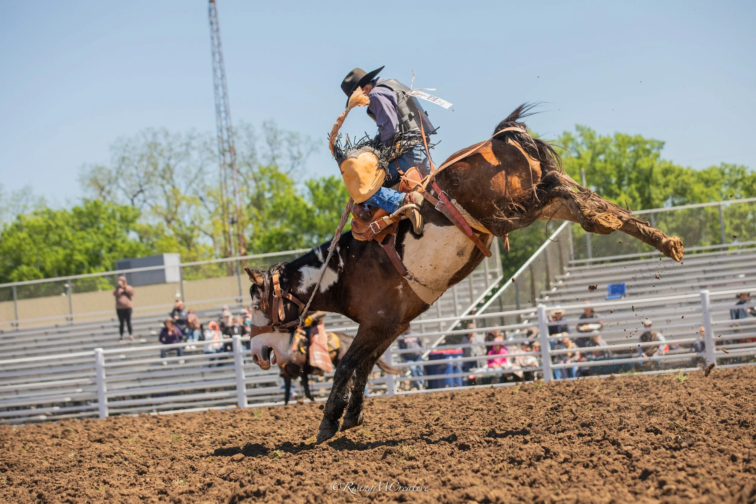 A cowboy wearing a hat and vest riding a bucking horse during a rodeo event, with spectators watching from the bleachers in the background.