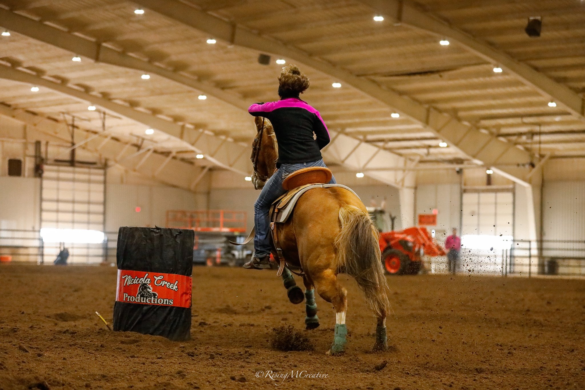 A person riding a horse inside an indoor arena, navigating around a black barrel marked with a red and white sign for Niccola Creek Productions, with a dirt-floor surface and a high arched ceiling.