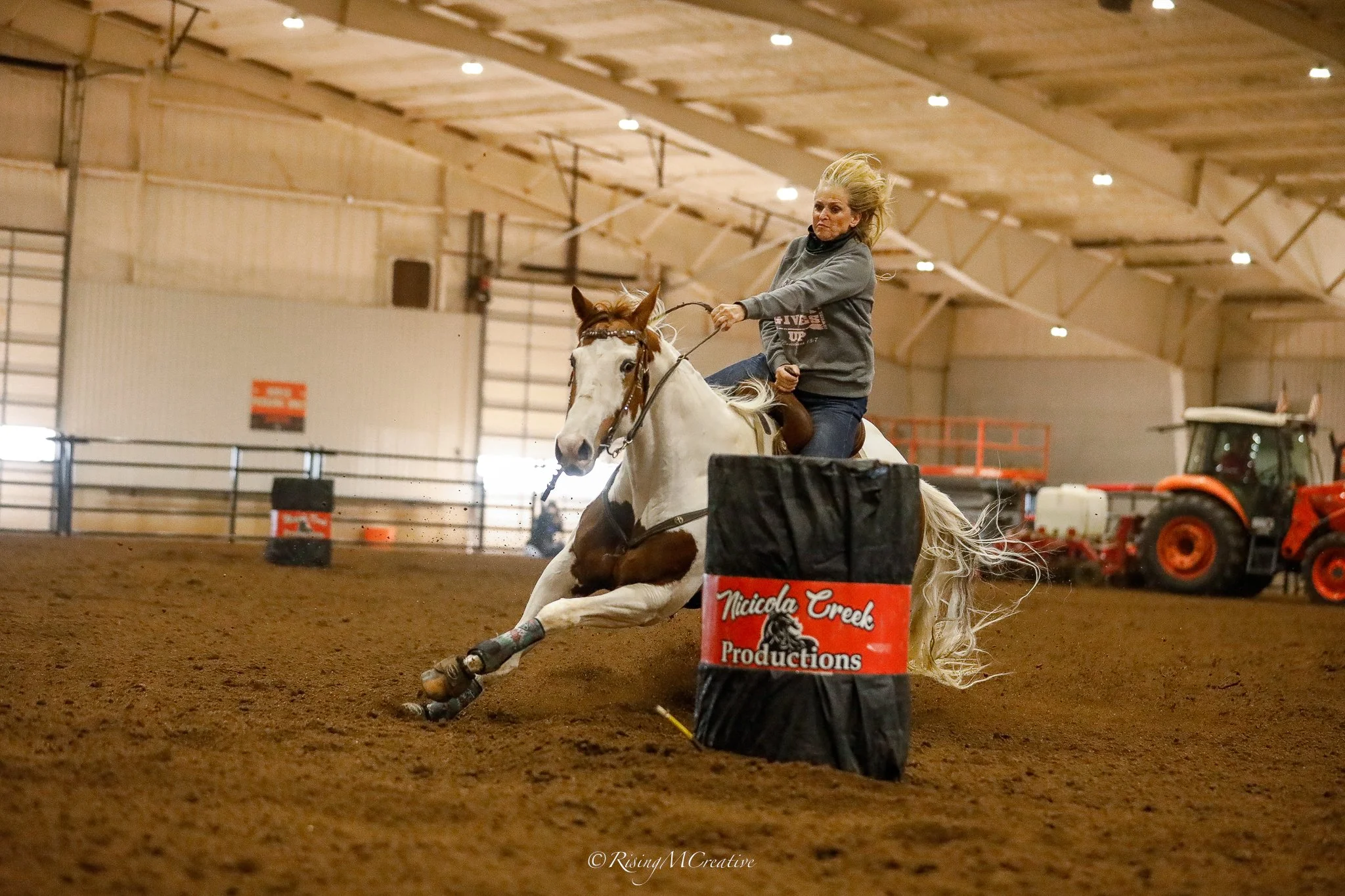A woman riding a horse in an indoor arena, participating in a barrel racing event, with orange cones and barriers around the course, and a tractor in the background.