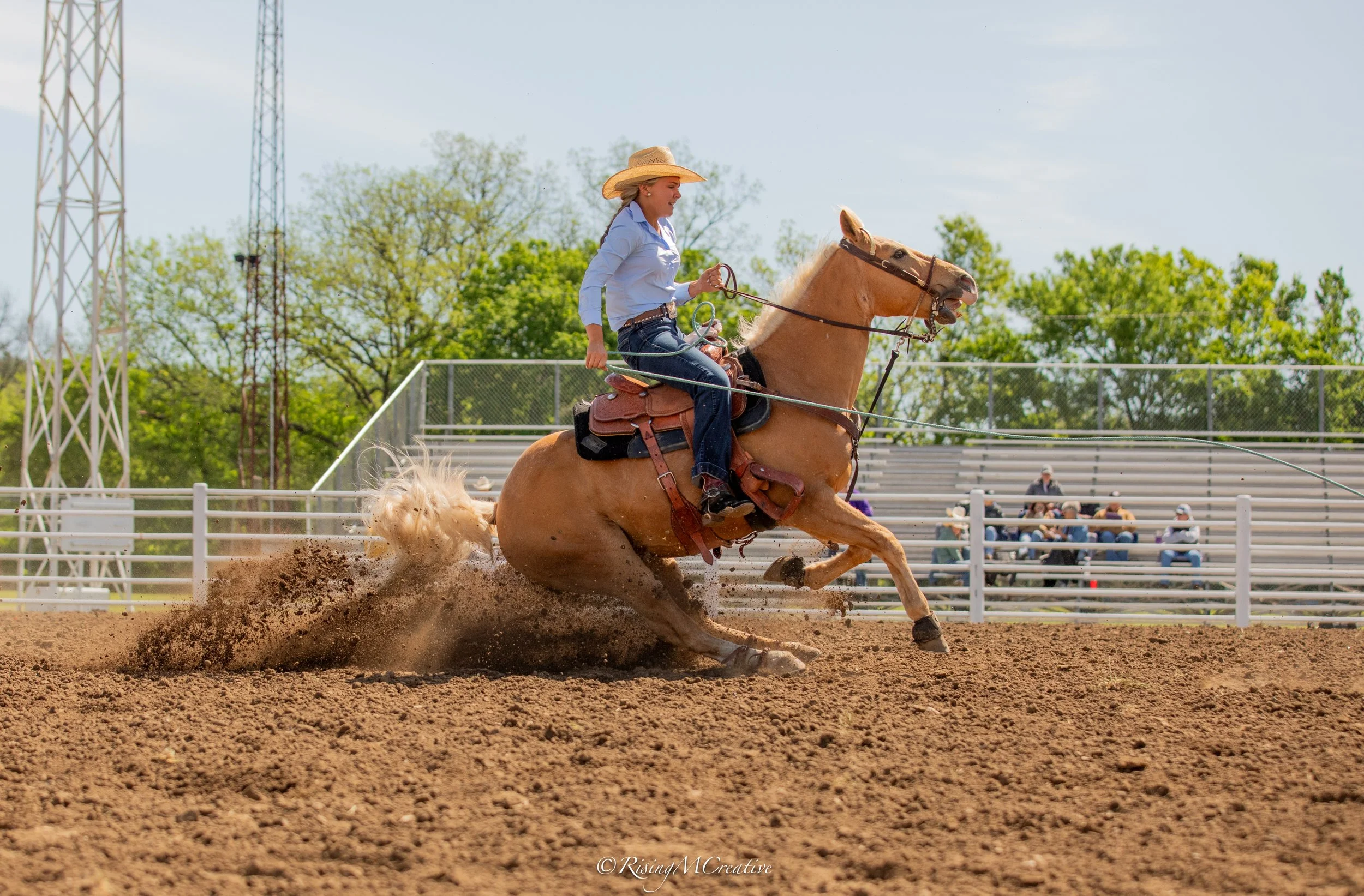A woman wearing a cowboy hat and light blue shirt riding a bucking horse in a rodeo arena with spectators and green trees in the background.