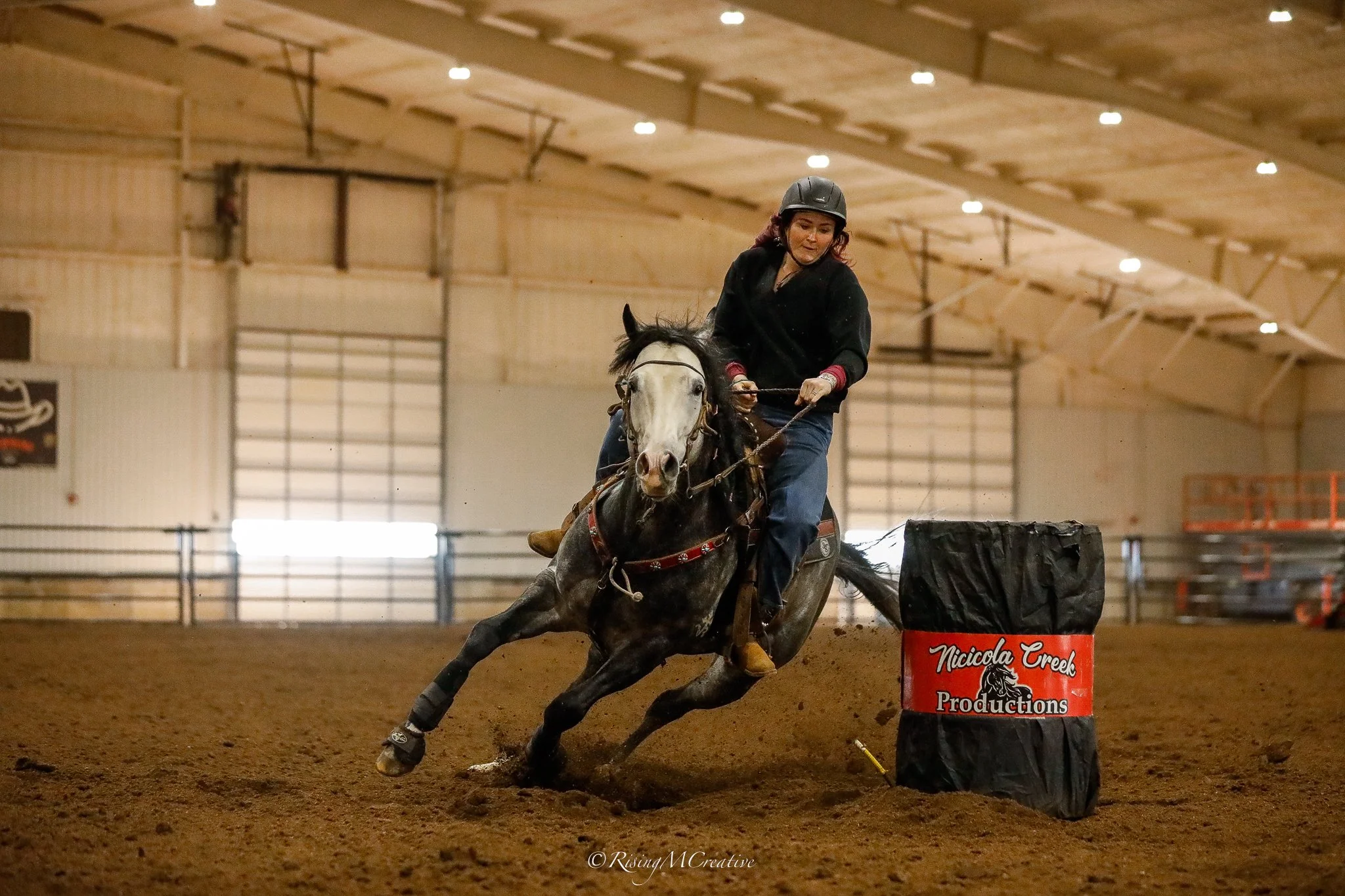A woman wearing a helmet and black jacket riding a horse through an indoor arena, navigating a barrel racing course. The arena has a wooden ceiling and a dirt floor, with a barrel labeled 'Nicciola Creek Productions.'