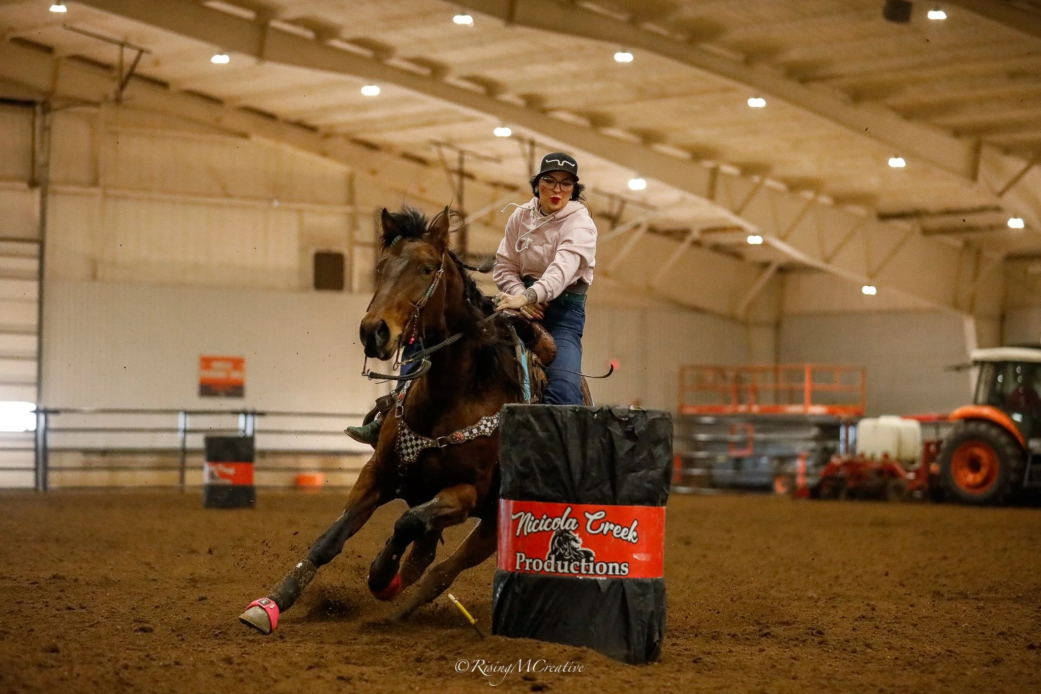 A woman riding a horse and jumping over an obstacle in an indoor equestrian arena. The woman is wearing glasses, a helmet, a light jacket, and jeans. The arena has a high ceiling with bright overhead lights, and there is a sign that reads 'Nicolcola 