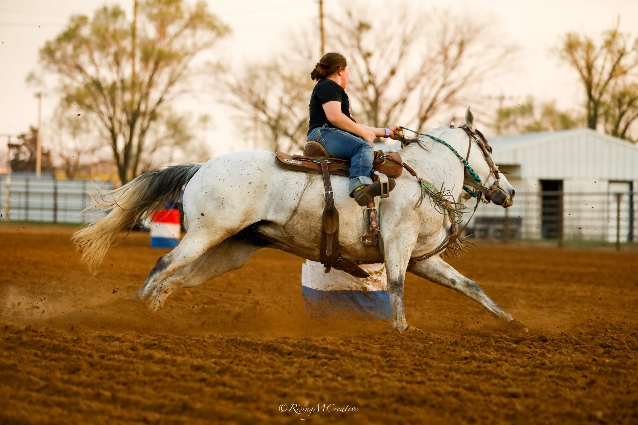 A person riding a white horse in an outdoor arena, with a dirt surface and a background of trees and a building, during a horse racing or riding event.