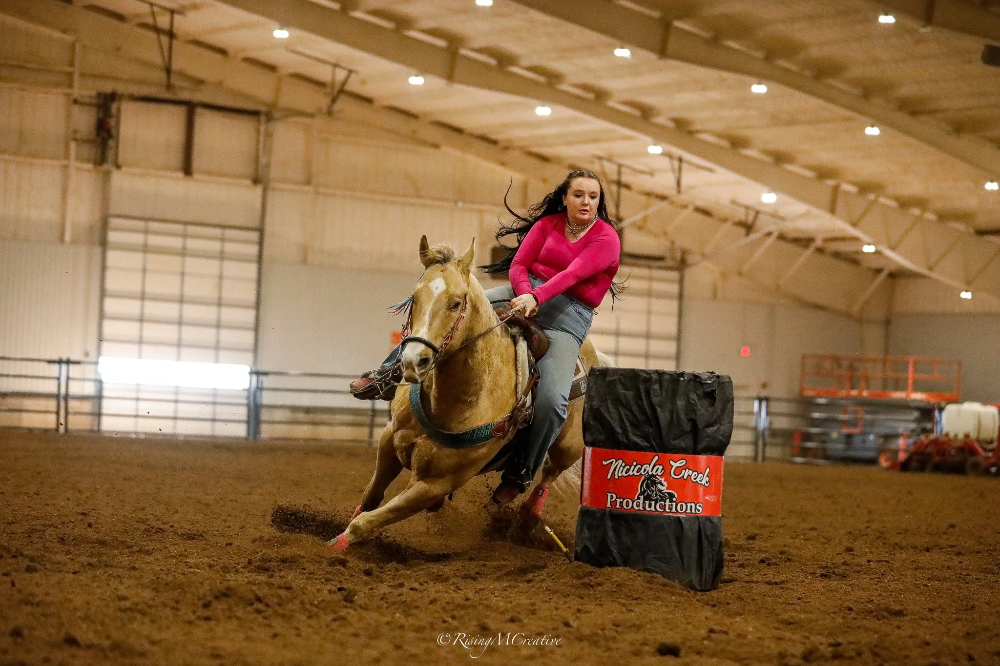A woman with long dark hair, wearing a bright pink long sleeve shirt, and gray jeans, is riding a light brown horse in an indoor arena. The horse is jumping over a barrel labeled 'Nicola Creek Productions'. The arena has a high wooden ceiling and met
