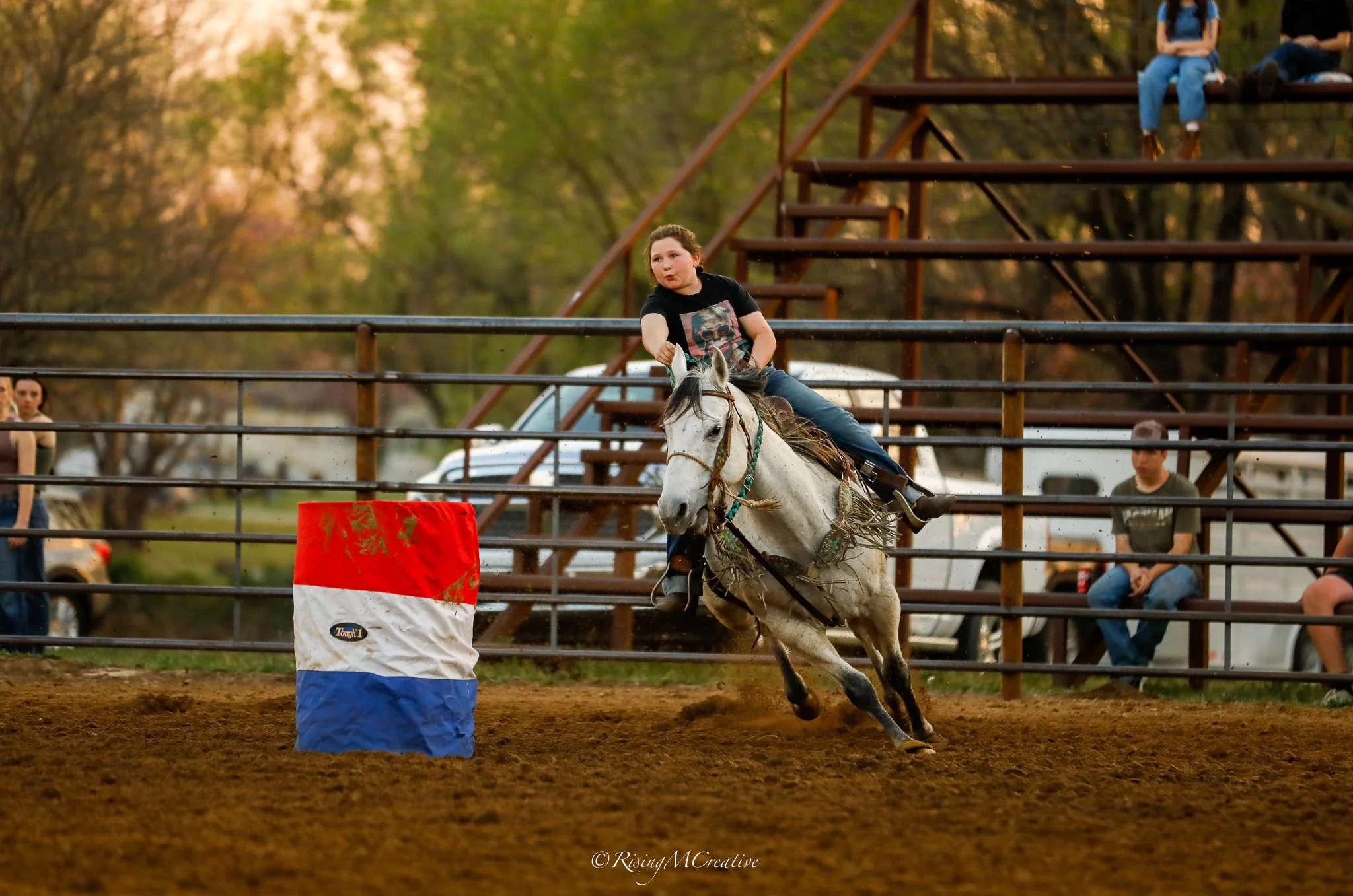 A girl riding a white horse on a dirt track during a rodeo, navigating around a barrel with red, white, and blue colors, with spectators and a wooden grandstand in the background.