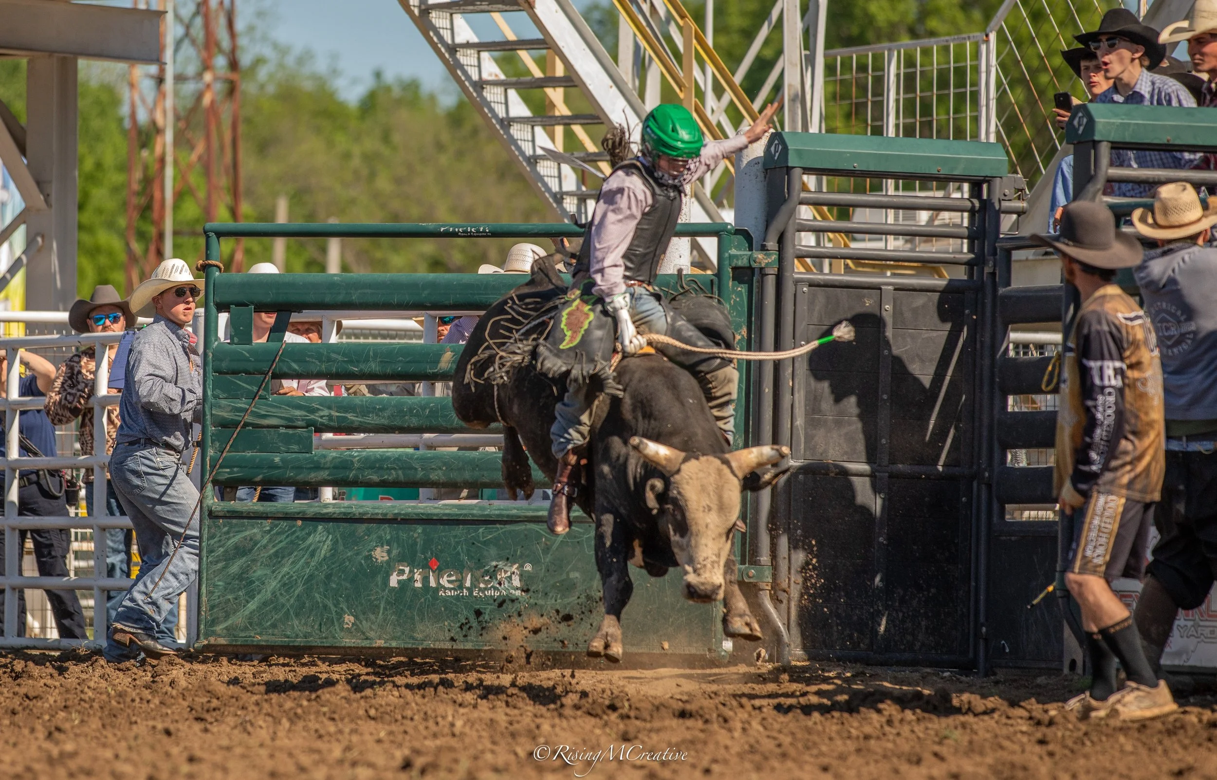 A cowgirl in a green helmet and cowboy attire riding a bucking bull during rodeo, with spectators and rodeo officials watching.