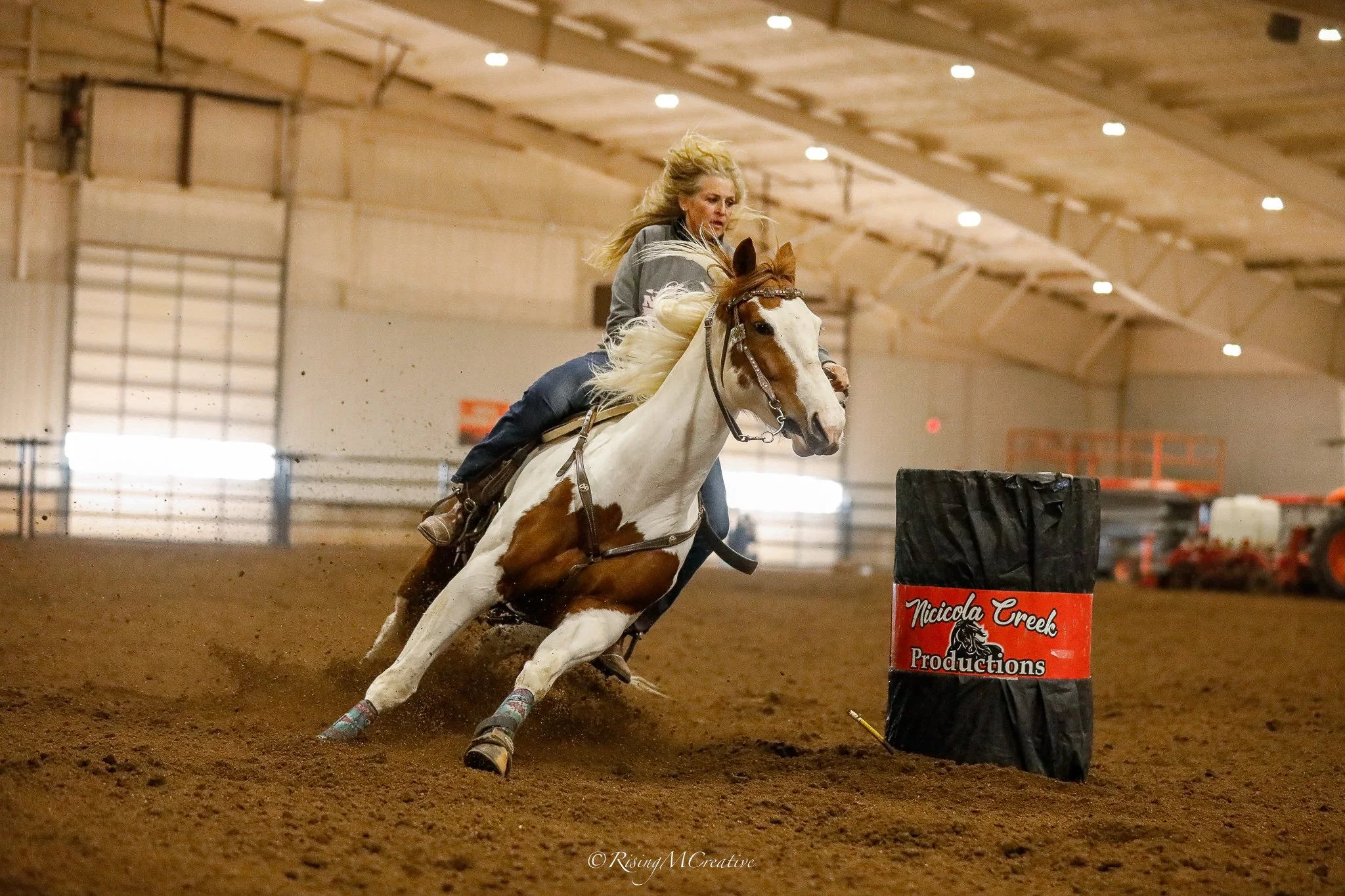 A woman riding a paint horse in an indoor rodeo arena, navigating around a barrel labeled 'Nicciola Creek Productions' during a barrel racing event.
