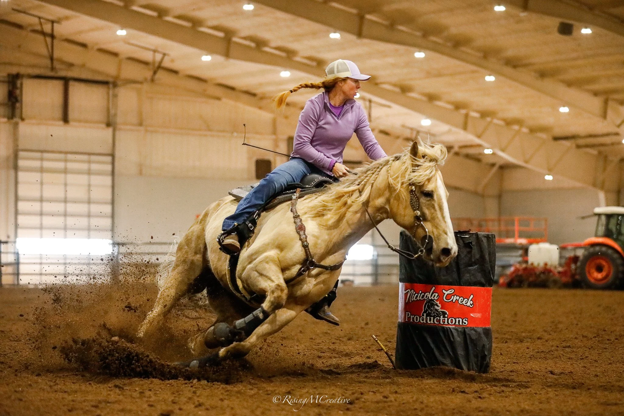 A woman riding a palomino horse in an indoor arena during a barrel racing event, guiding the horse around a barrel marked 'Nicola Creek Productions'.