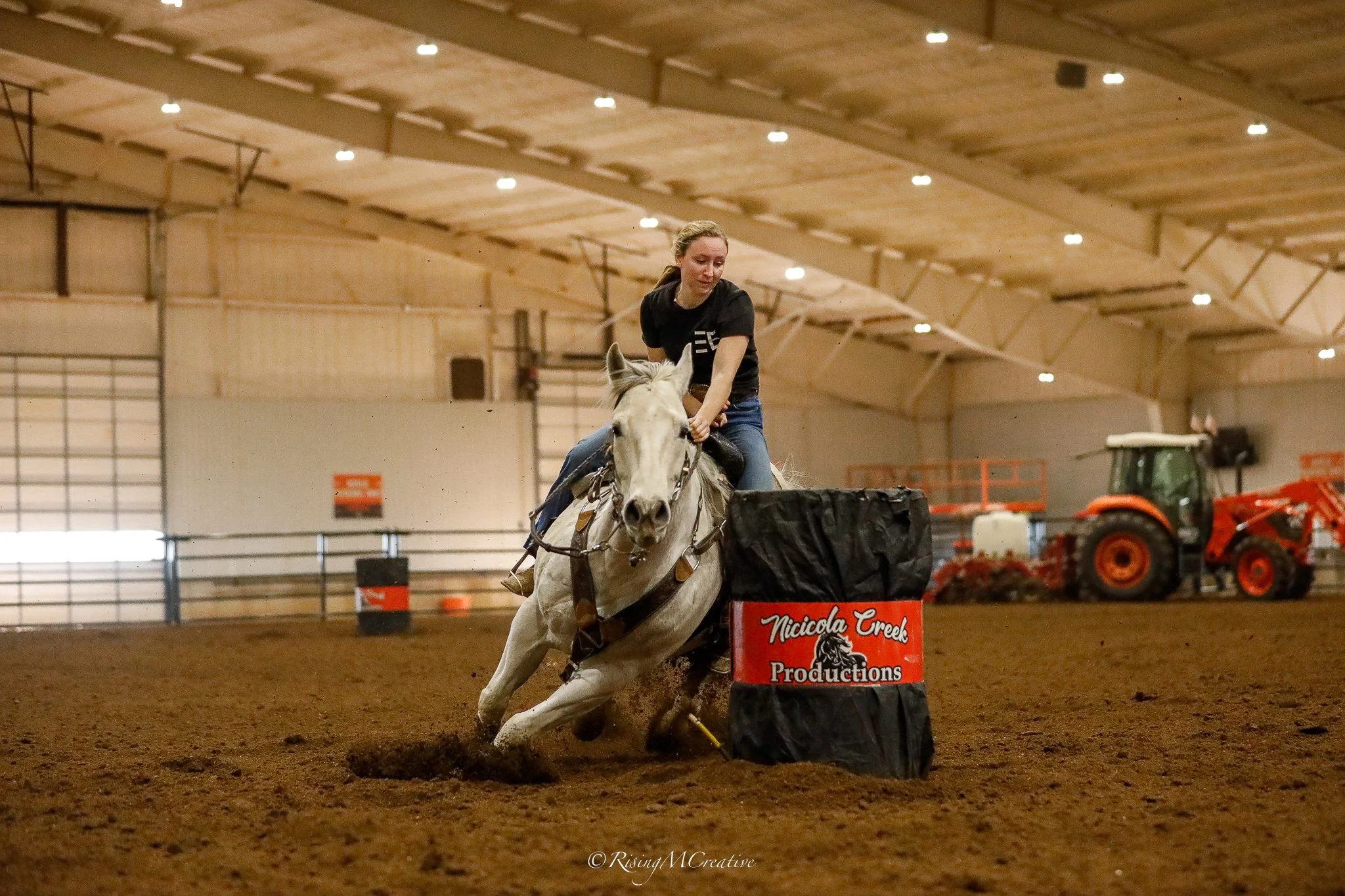 A woman riding a white horse in an indoor arena, participating in a barrel racing event. The horse is maneuvering around a barrel labeled 'Nicolica Creek Productions.' In the background, farm equipment such as a tractor and other machinery are visibl