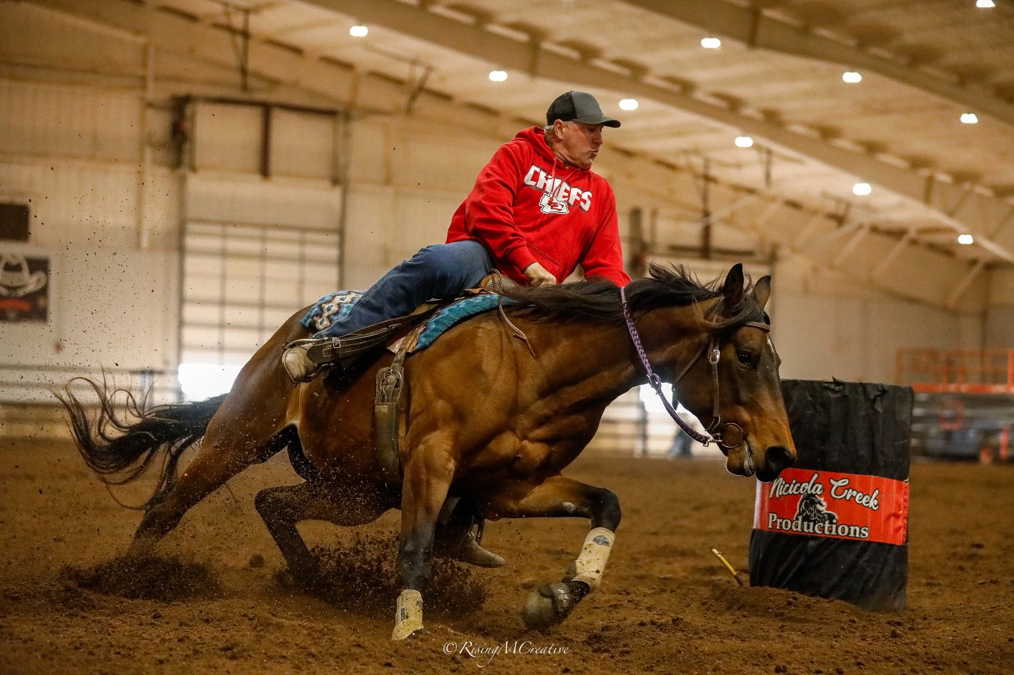 A person wearing a gray and black cap and a red hoodie with the word 'Chiefs' riding a galloping brown horse in an indoor arena, dust and dirt flying behind them.