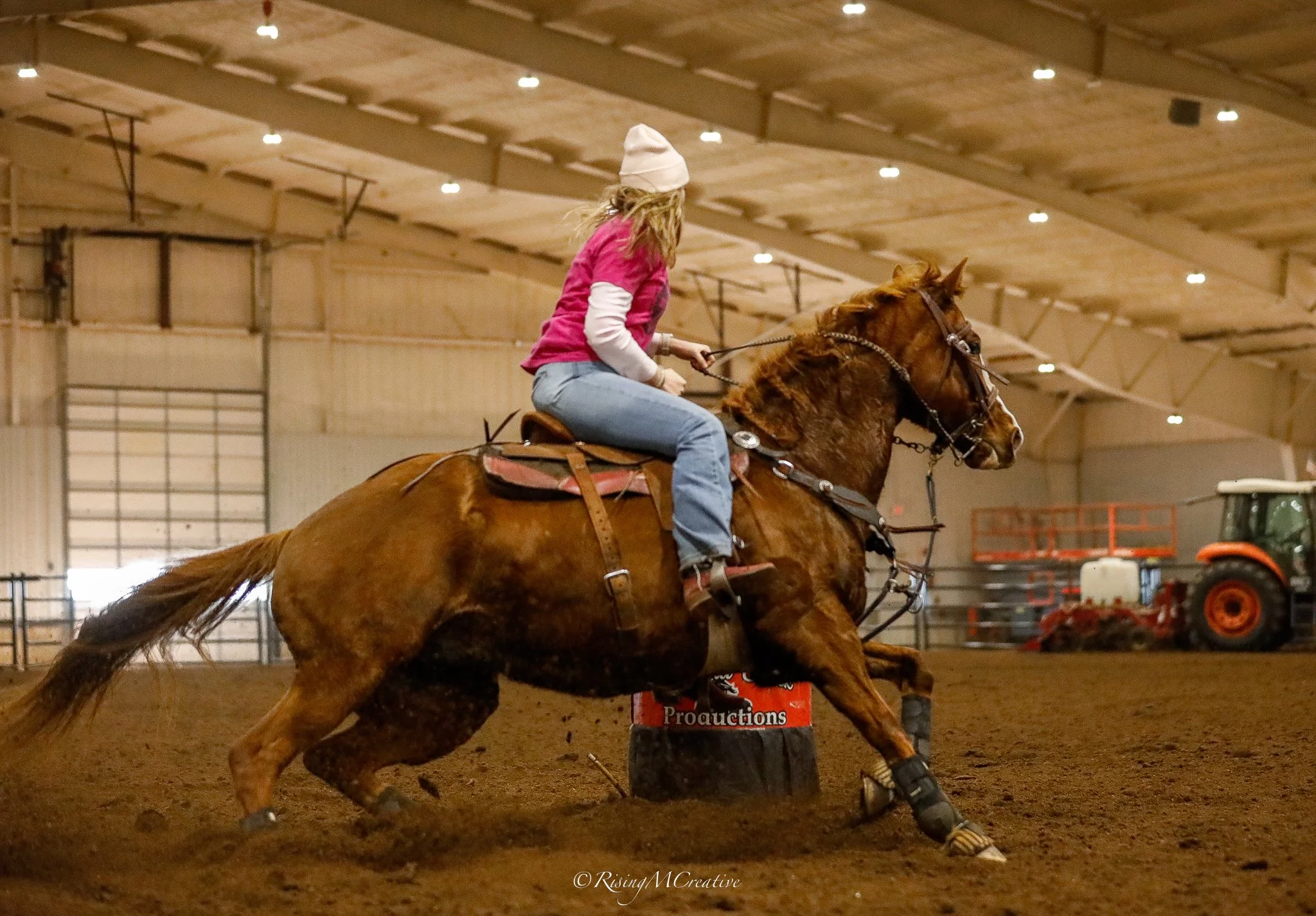 A rider in a pink shirt and white hat competing in an indoor equestrian event, riding a galloping brown horse.