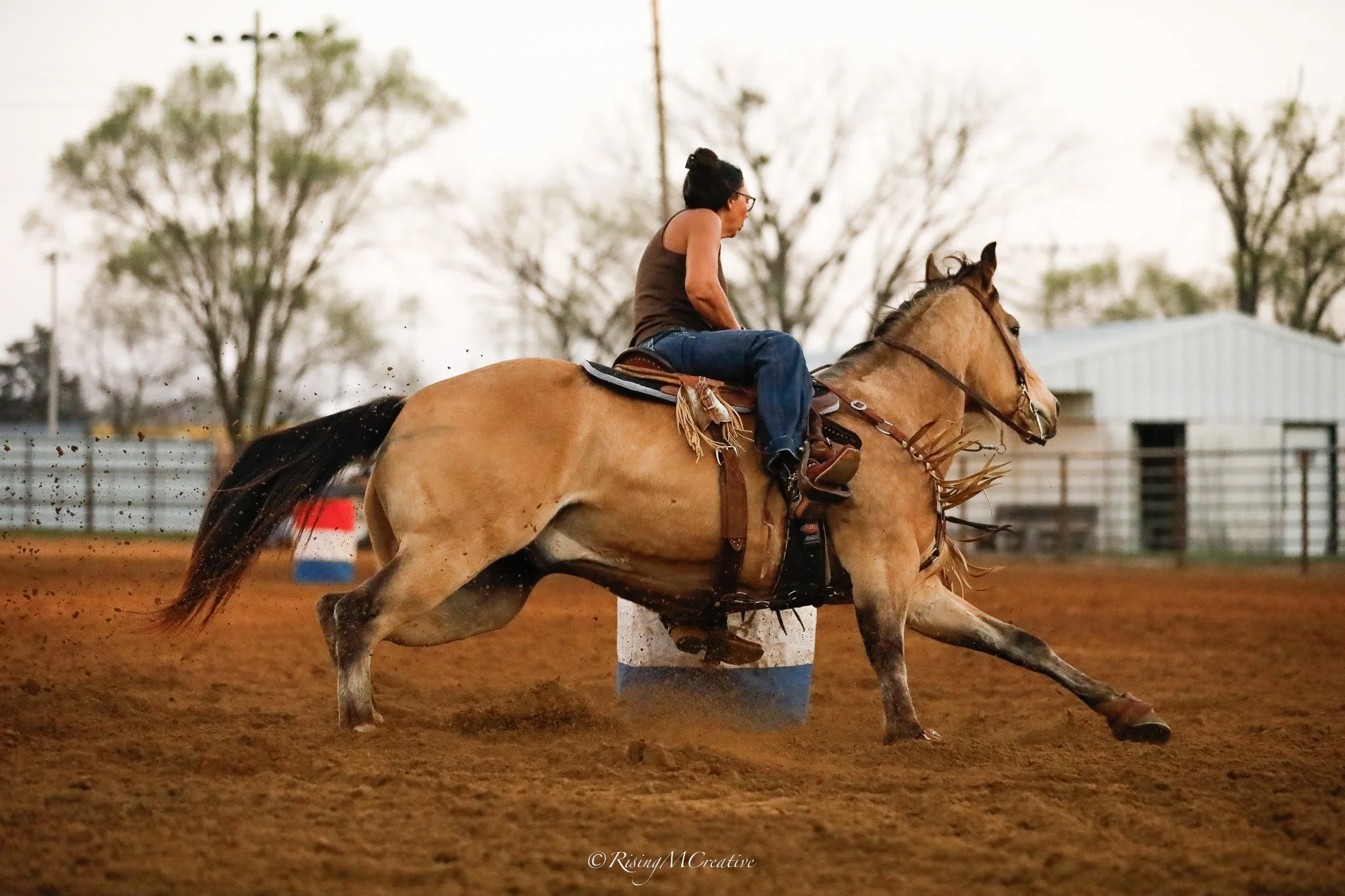 A woman riding a bucking horse inside an arena, dirt flying from the horse's hooves, with trees and a building in the background.