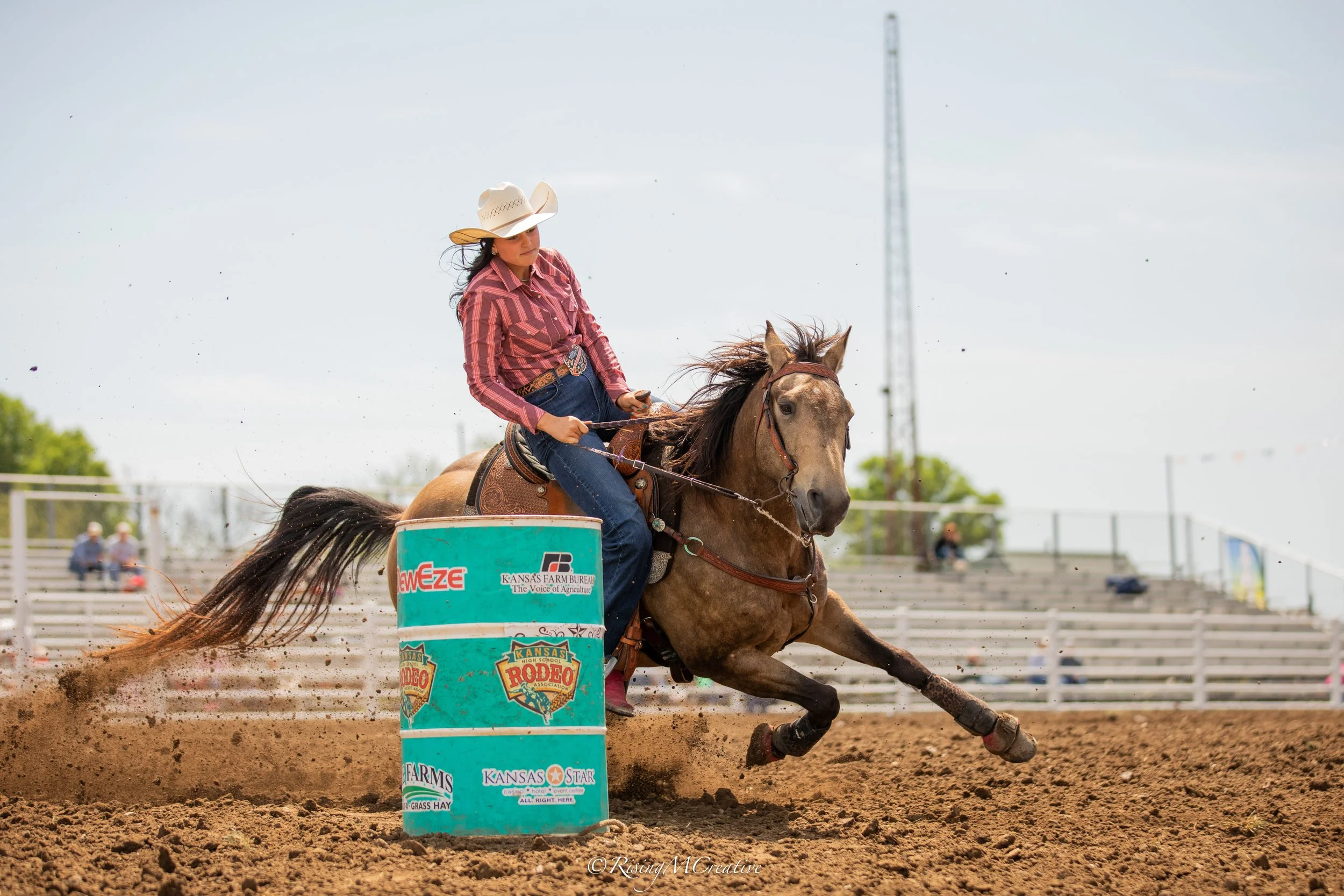 A female rodeo rider in a plaid shirt and cowboy hat riding a bucking horse around a barrel on a dirt arena, with a fence and spectators in the background.