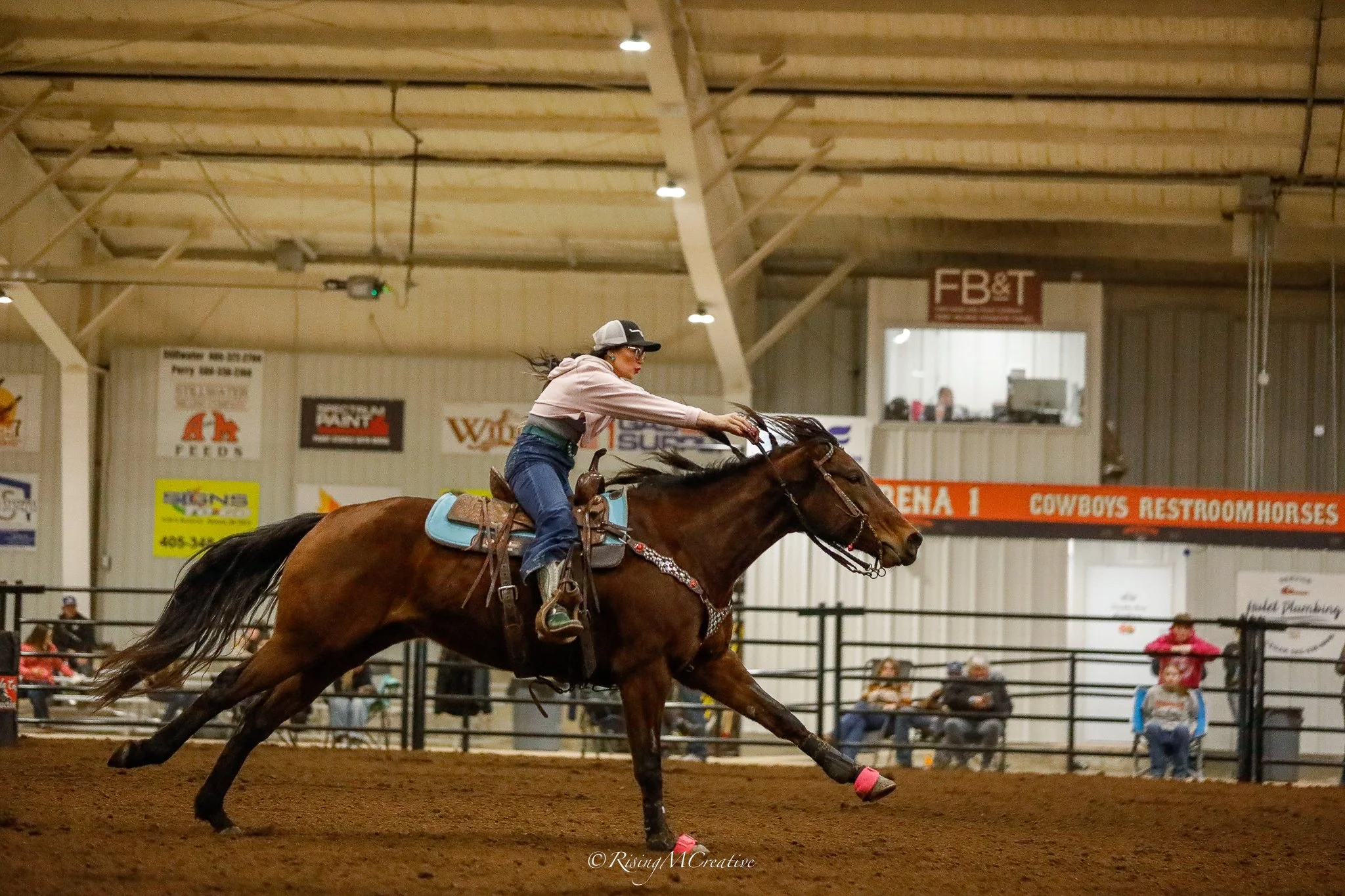 A woman wearing a cap and glasses riding a galloping horse in an indoor arena, with spectators and banners in the background.