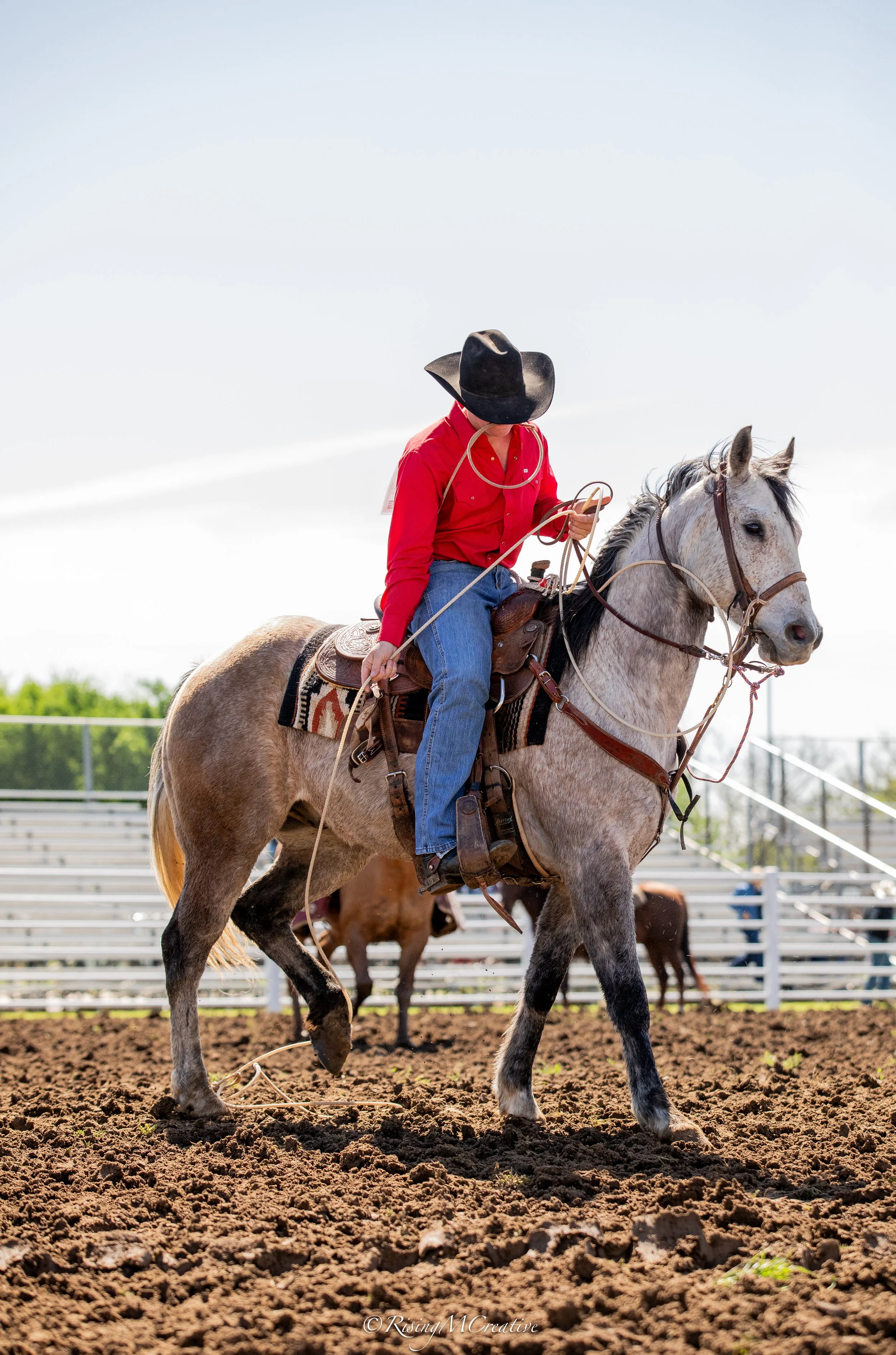 A cowboy wearing a red shirt, jeans, and a black hat riding a gray horse on a dirt arena with a metal fence and trees in the background.