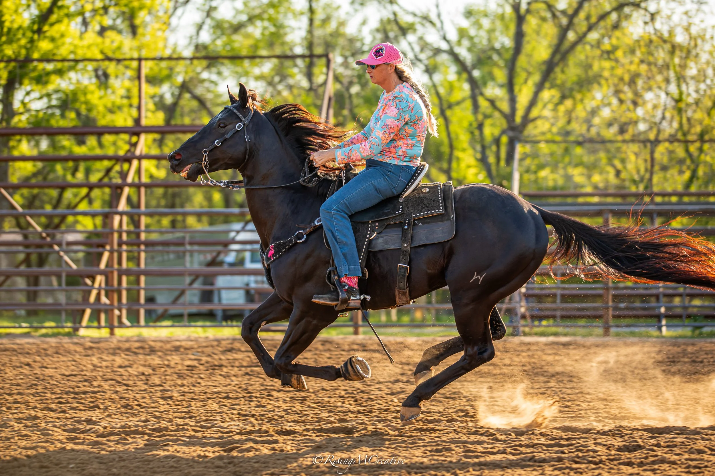 A woman riding a black horse on a dirt riding arena, with green trees in the background, during the daytime.