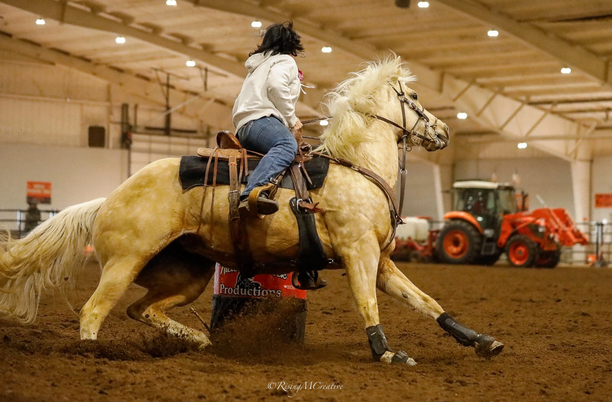 A person riding a bucking palomino horse inside an indoor arena with tractor equipment in the background.