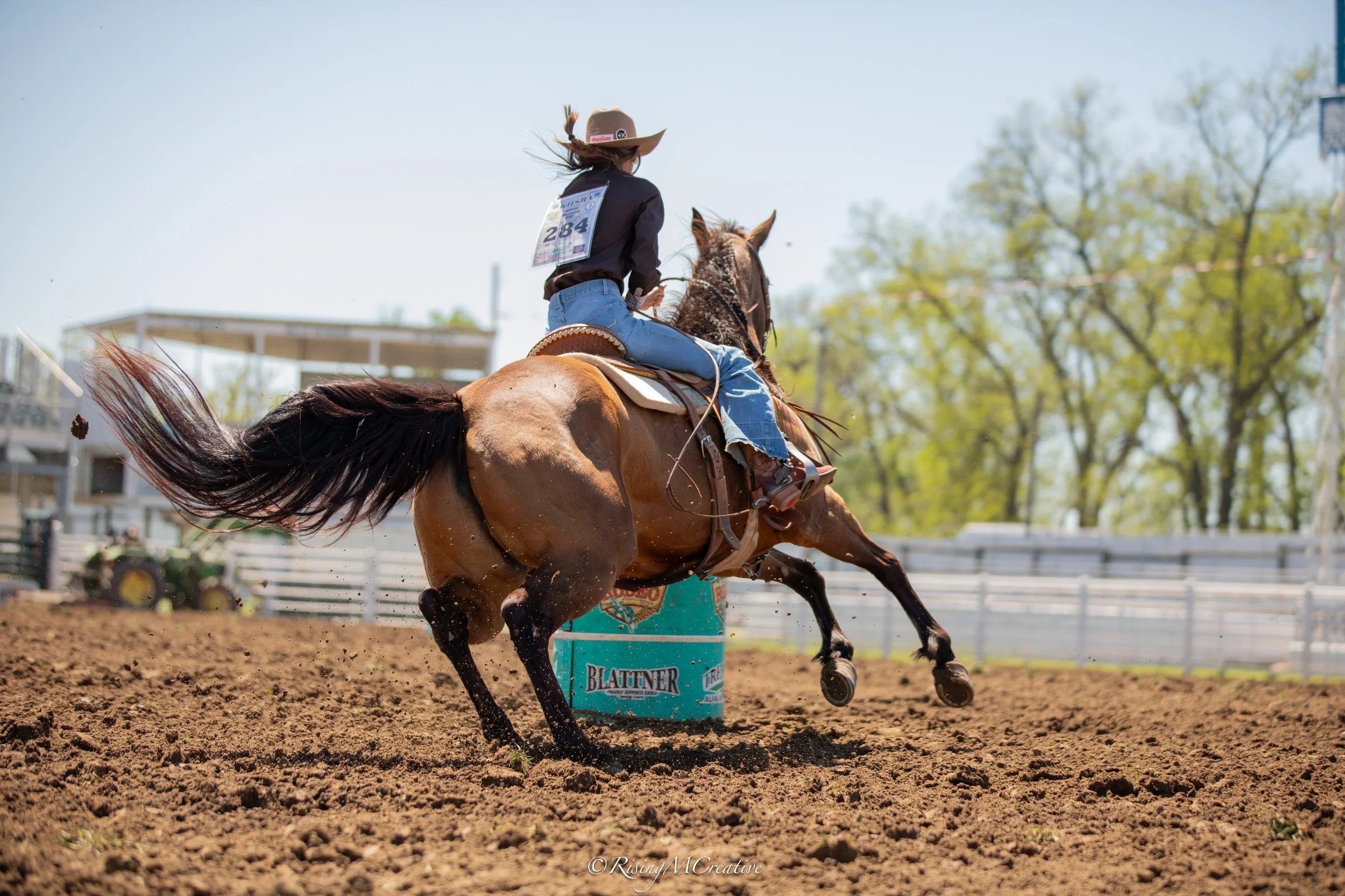 A rodeo rider on a bucking horse during a competition, with a barrel marked 'BLATTNER' in the dirt arena, trees in the background, and a clear sky.