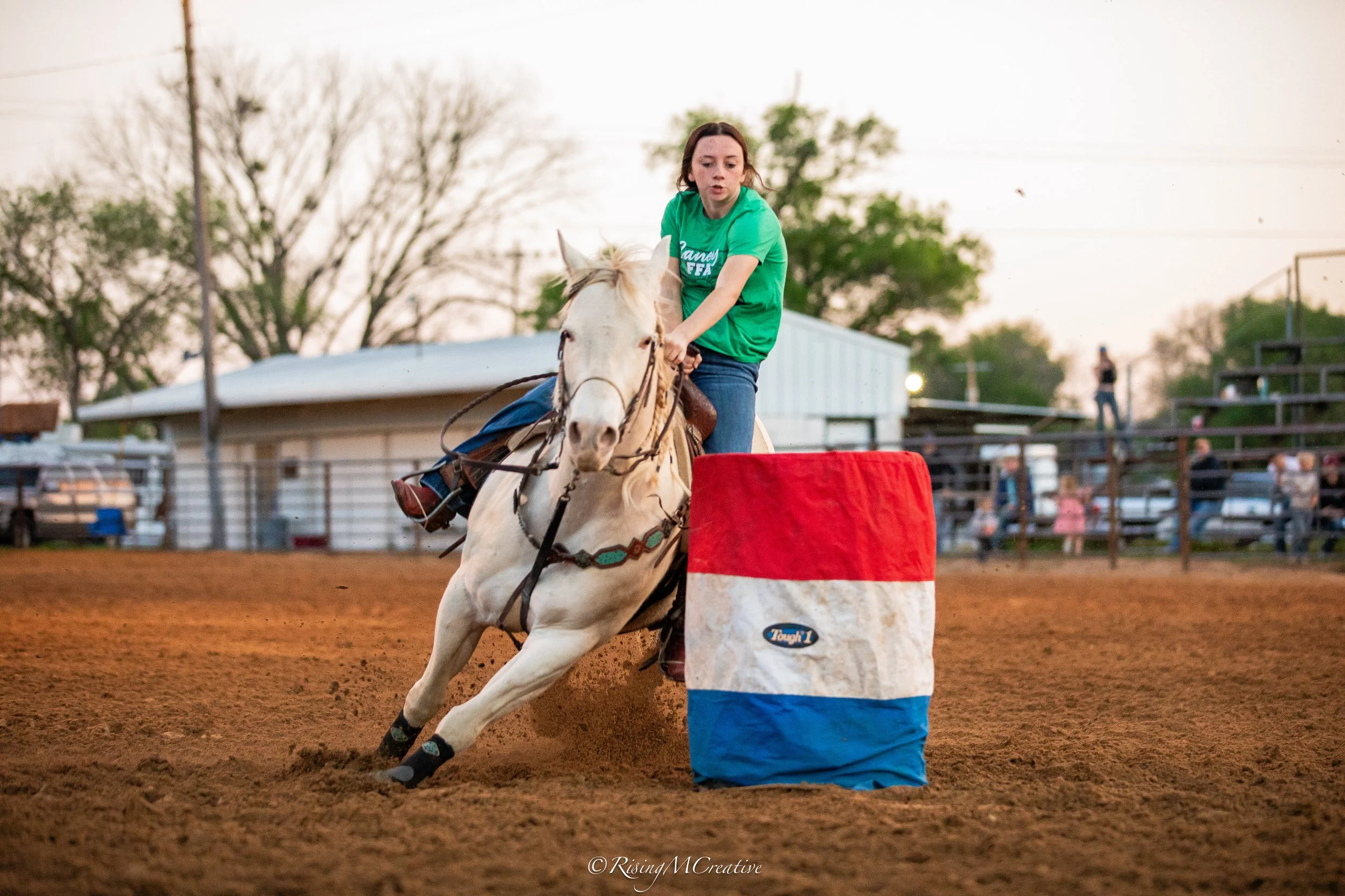 A woman wearing a green shirt riding a white horse during a barrel racing event at an outdoor arena with trees and spectators in the background.