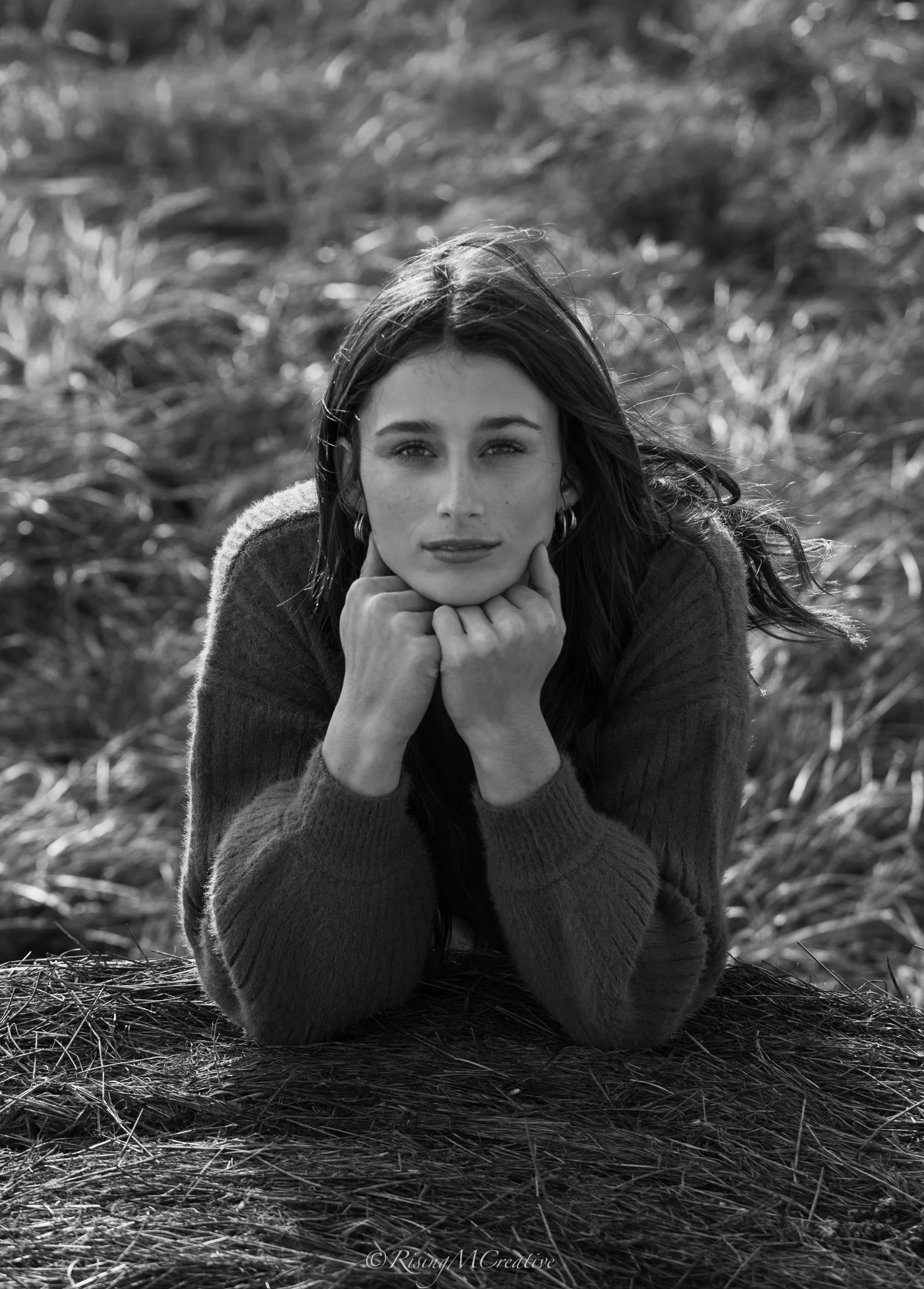 Black and white photo of a woman lying on her stomach on a grassy field, resting her chin on her hands, looking into the camera.