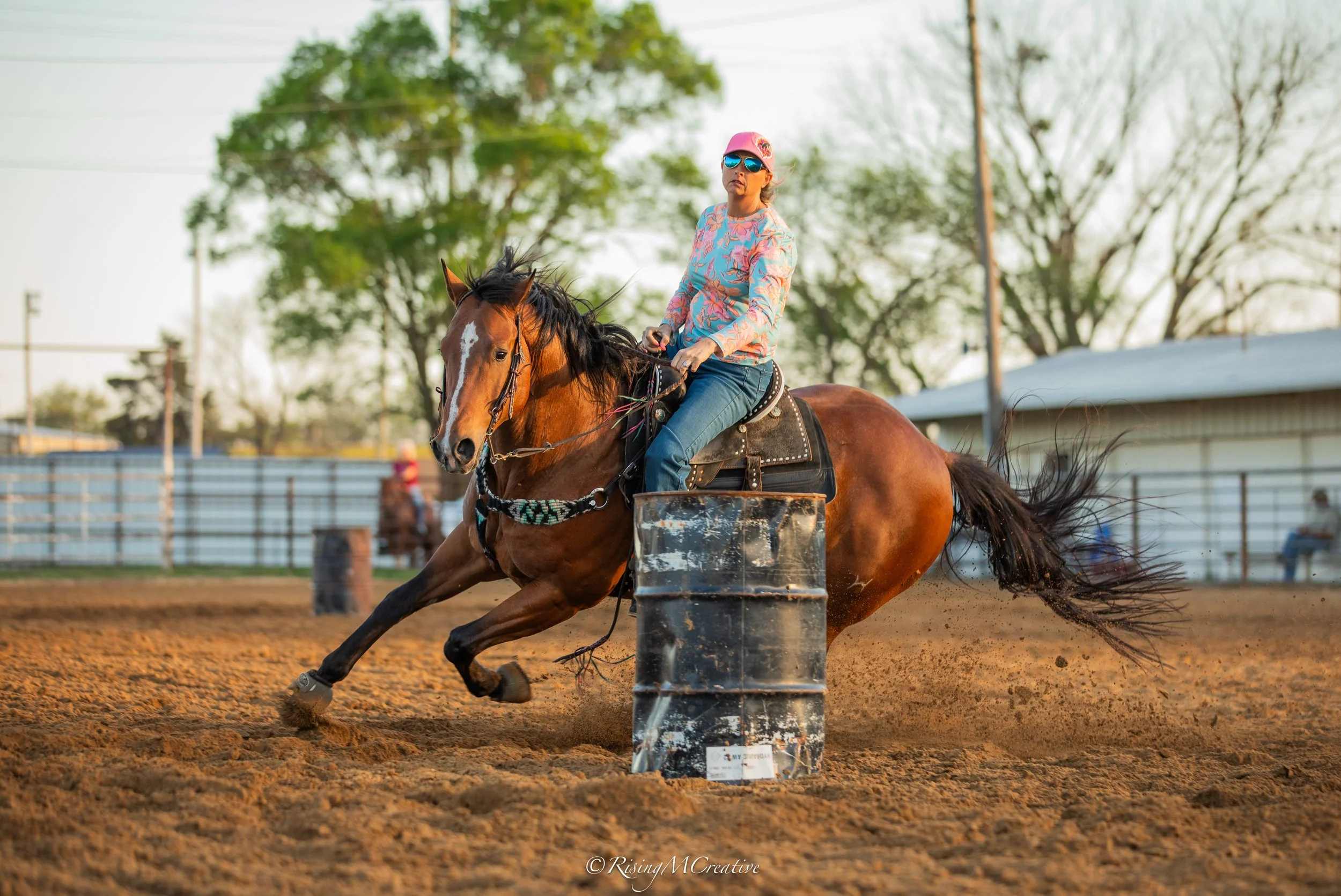 A woman riding a brown horse around a barrel at a rodeo or horse racing event during daytime. She is wearing sunglasses, a pink cap, a colorful long-sleeve shirt, and jeans. The photograph captures her in motion as she navigates the turn, with a back
