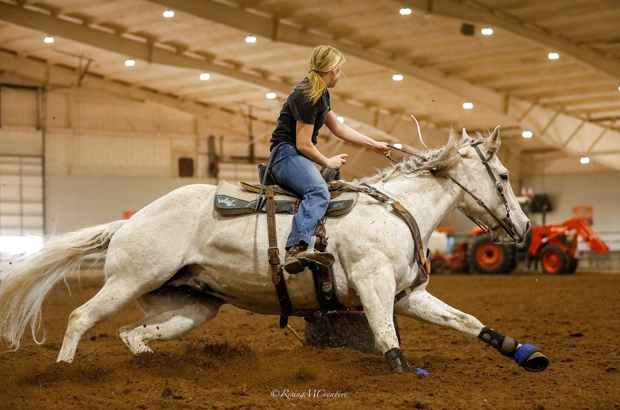 A woman riding a white horse in an indoor arena, participating in a rodeo event, with dirt floor and equipment in the background.