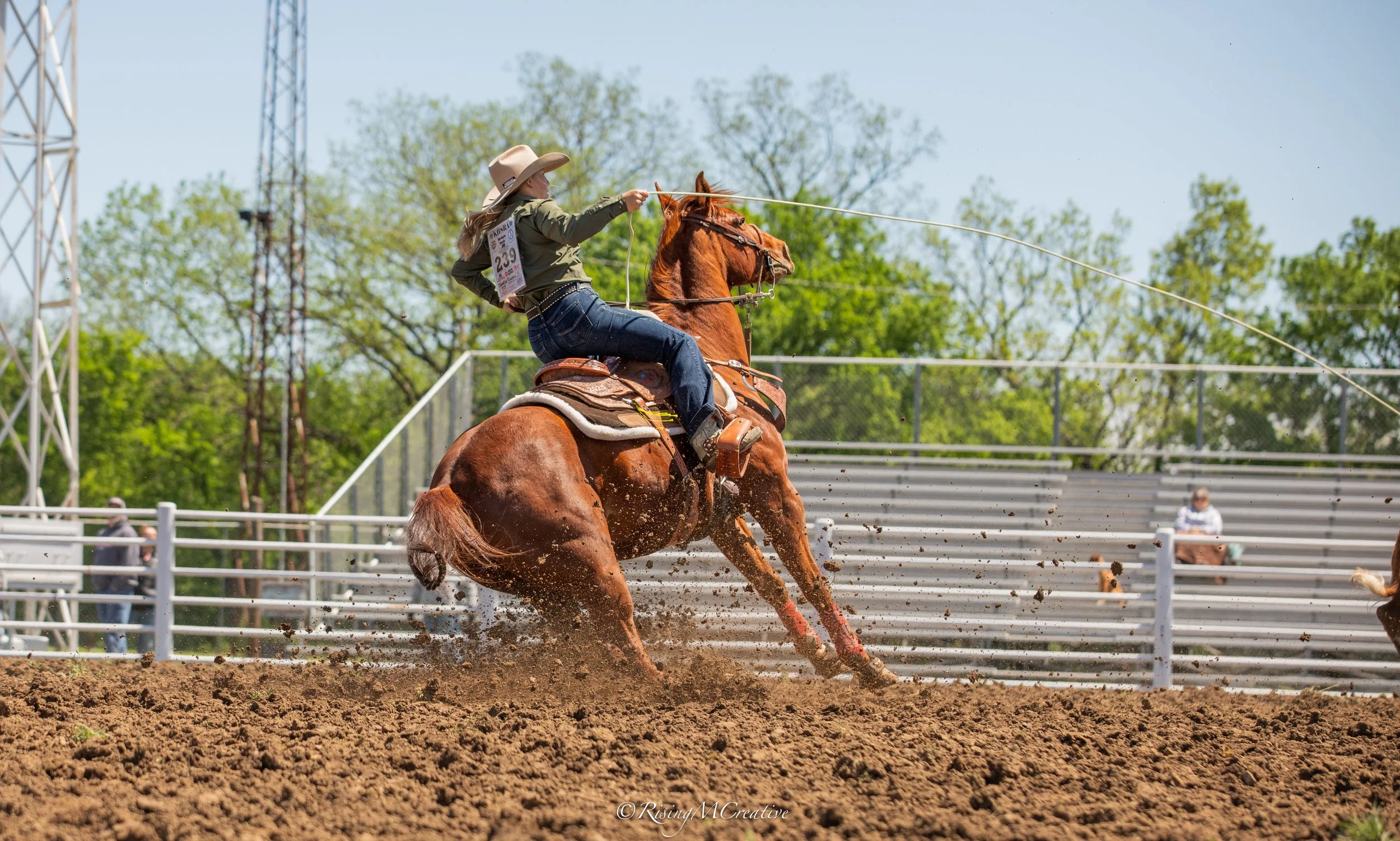 A woman wearing a cowboy hat and western-style attire riding a galloping brown horse in a rodeo arena, with spectators and green trees in the background during daytime.