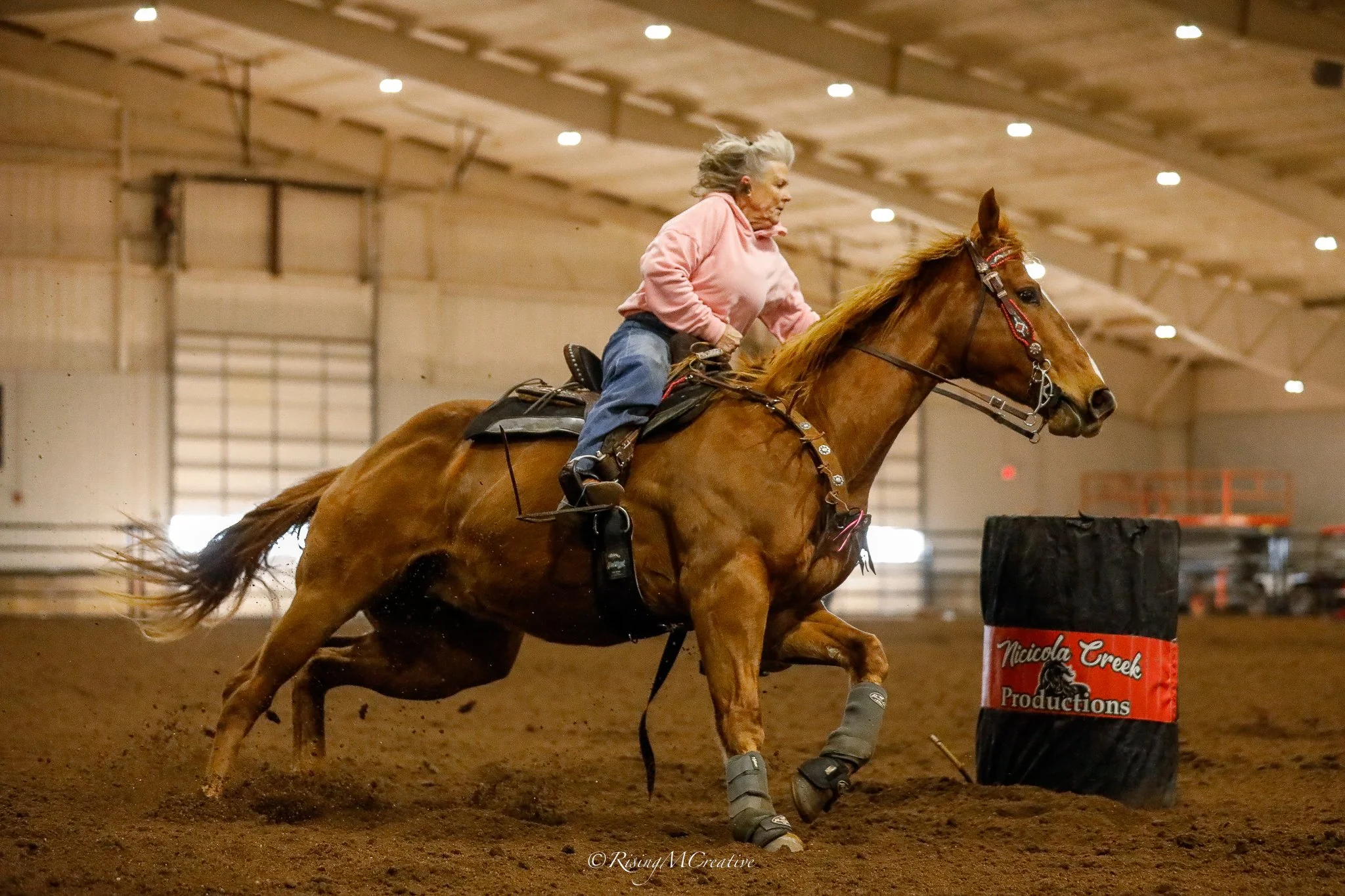 An elderly woman riding a horse indoors during a barrel racing event, navigating around a barrel with a focused expression.