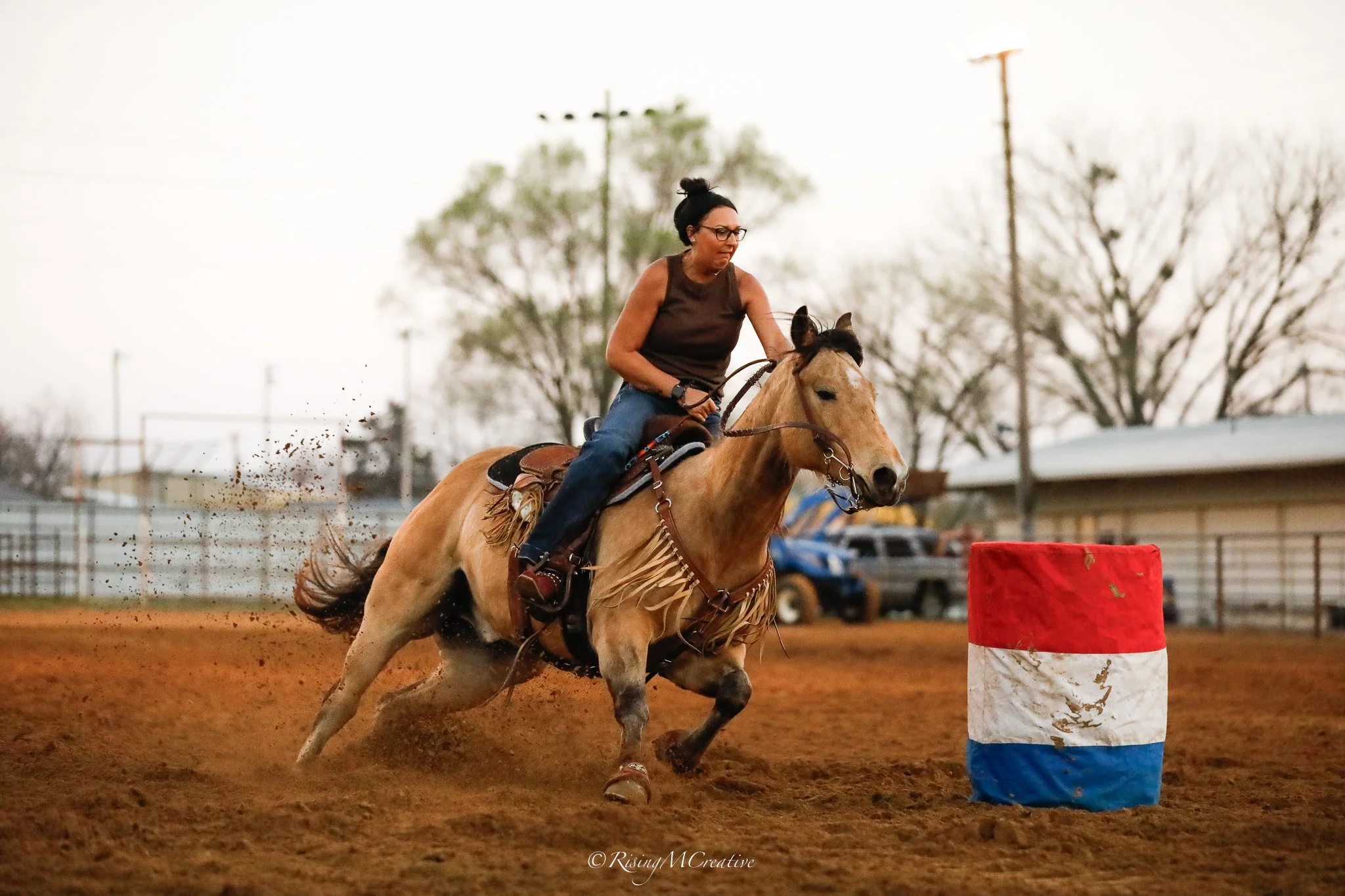 A woman riding a horse on a dirt racetrack during a rodeo or horse racing event. She is wearing glasses and a sleeveless top, and is focused on the race. The horse is galloping past a red barrel, with trees and some structures blurred in the backgrou