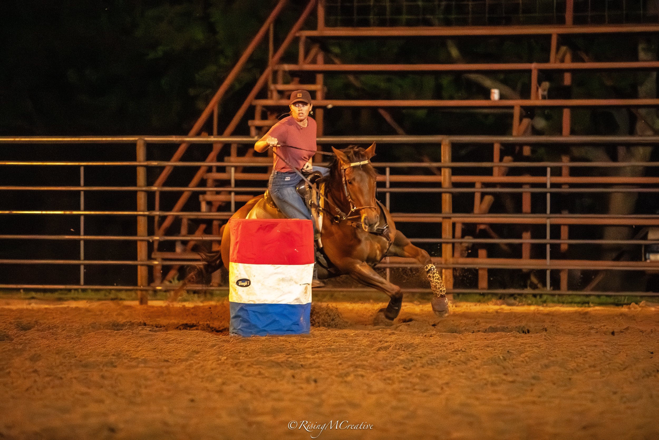 A person riding a brown horse during a nighttime equestrian event. The rider is wearing a maroon shirt, jeans, and a helmet, and appears to be navigating around a red and white barrel on a dirt arena with wooden bleachers in the background.