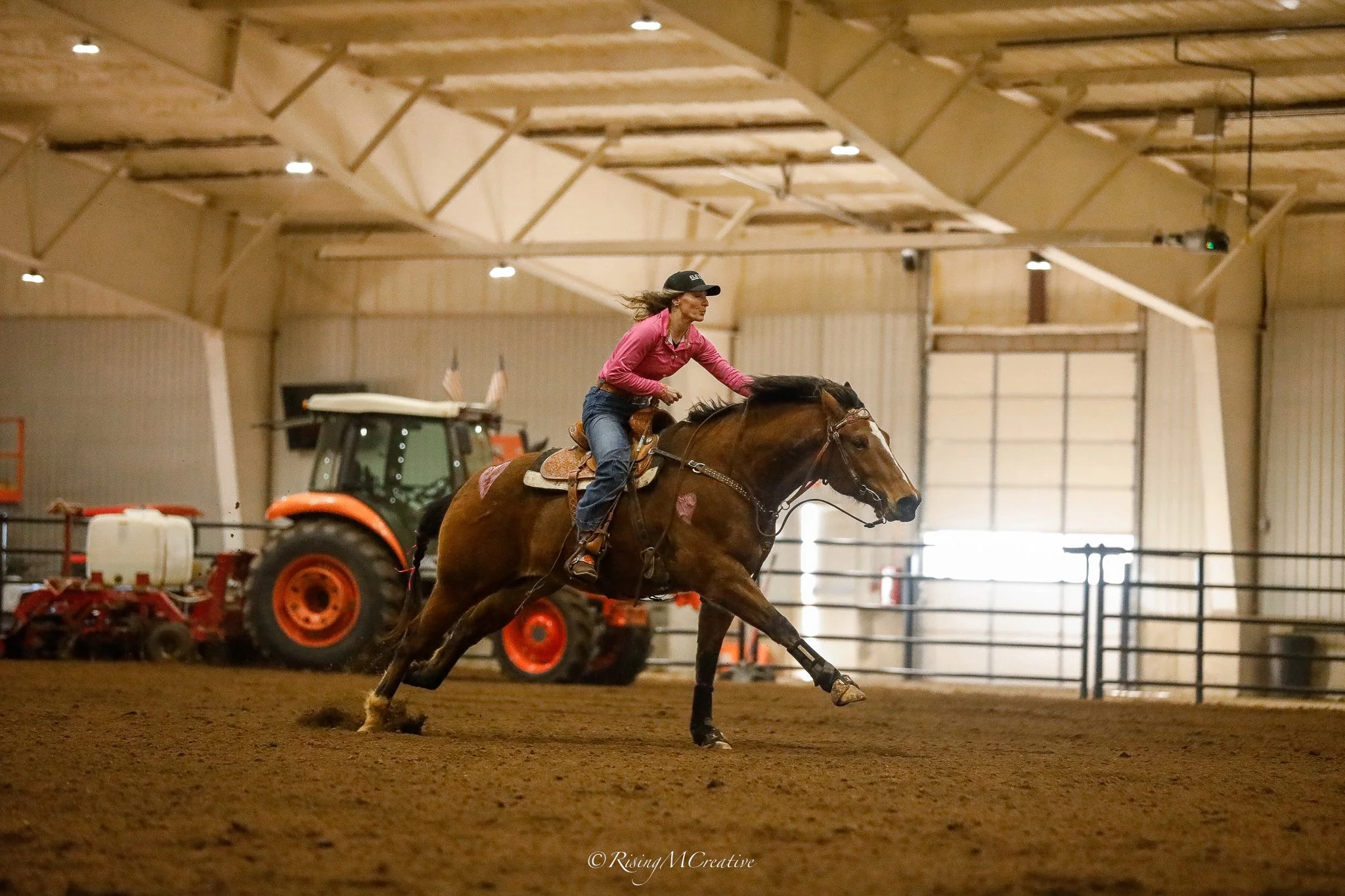 A woman riding a horse in an indoor arena, with farming equipment and an orange tractor in the background.