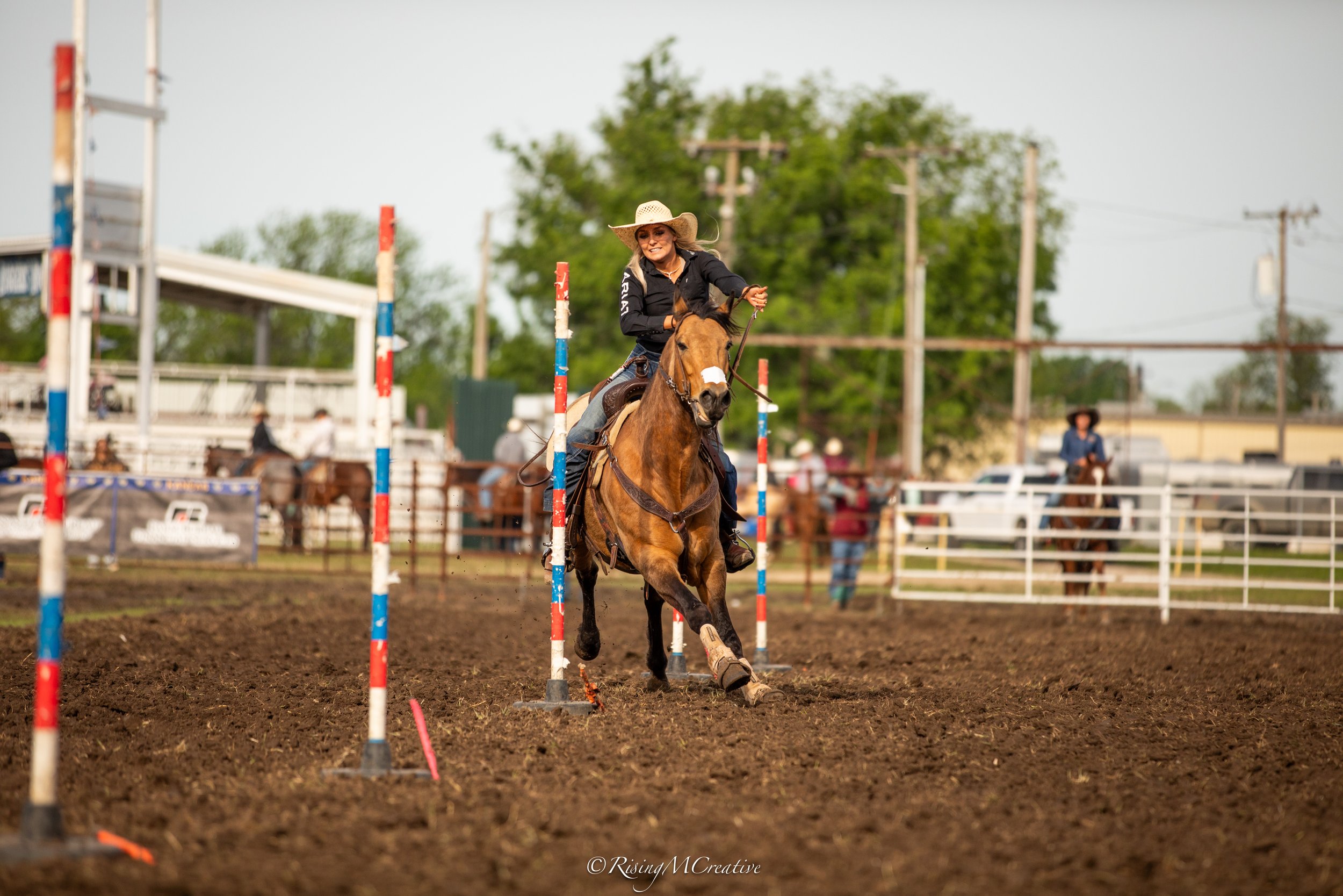 A woman wearing a white cowboy hat and black shirt rides a brown horse and participates in a barrel racing event at a rodeo arena, with support staff and other riders visible in the background.