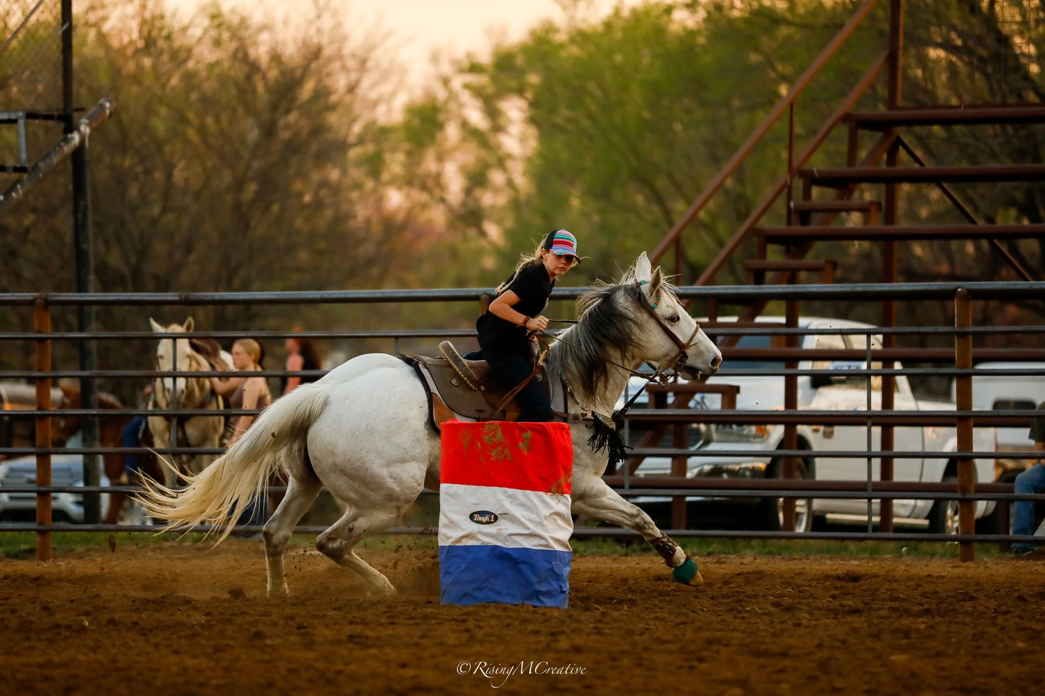 A girl riding a white horse in a fenced arena during a competition, with an orange and blue barrel in front. Trees and spectators are visible in the background, and the scene appears to be during sunset.