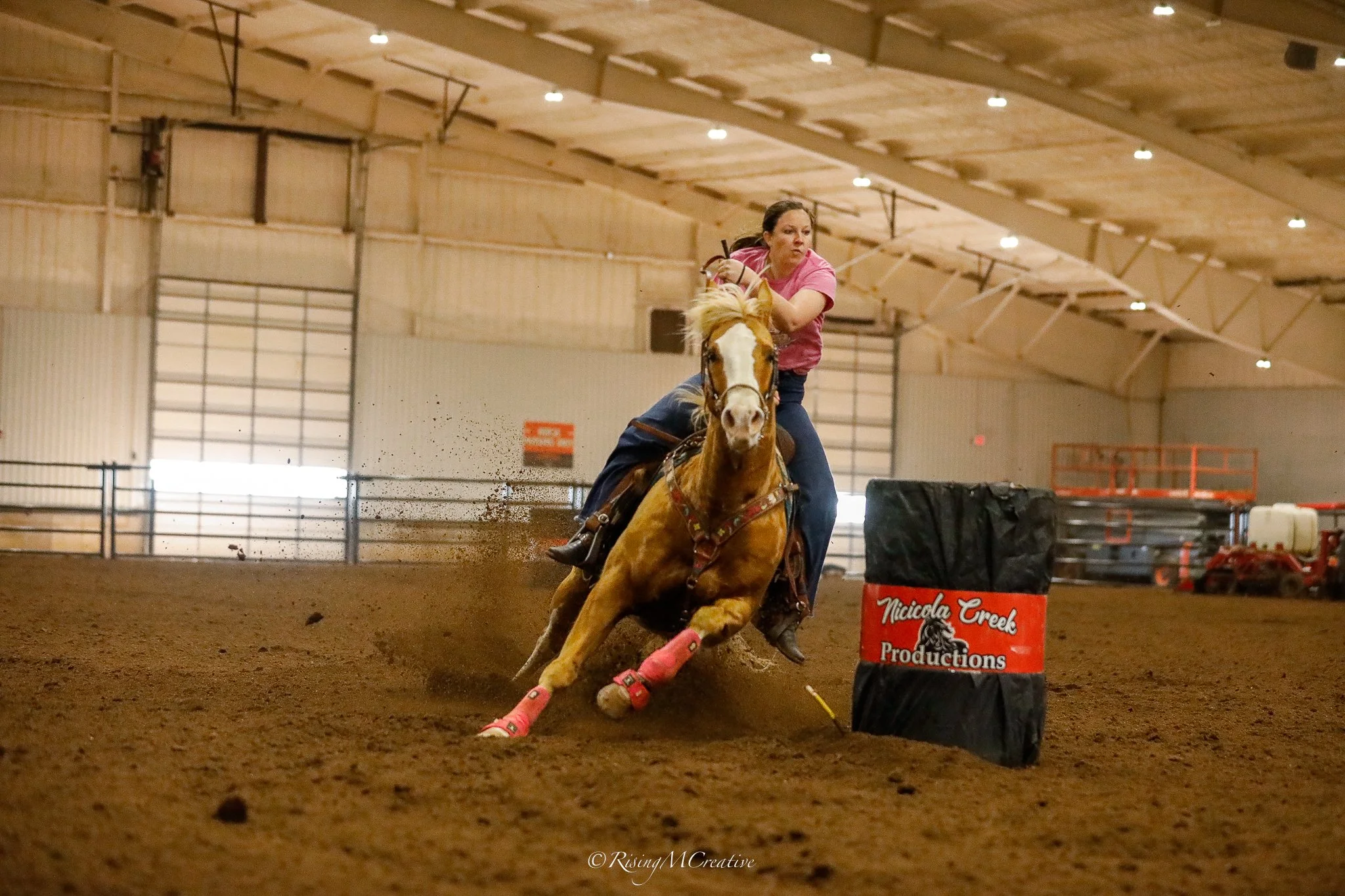 A woman riding a horse during a barrel racing event inside an indoor arena. She is wearing a pink shirt and jeans, and the horse is maneuvering around a barrel marked with "Nicolado Creek Productions." The arena has a high ceiling with lights and a d